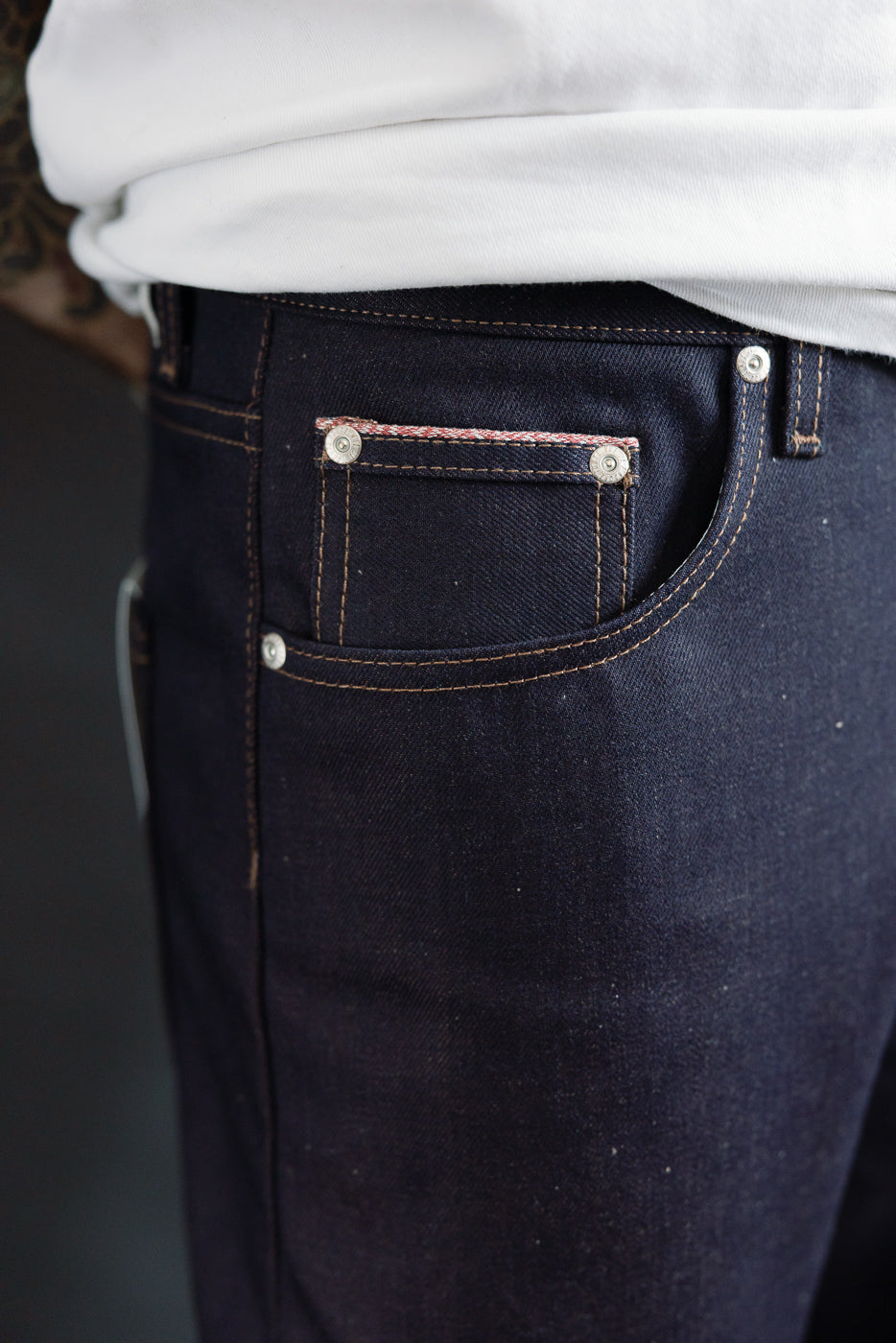Close-up of male model’s right front pocket and coin pocket on dark selvedge jeans, showing texture and stitching detail. Model is 5'9