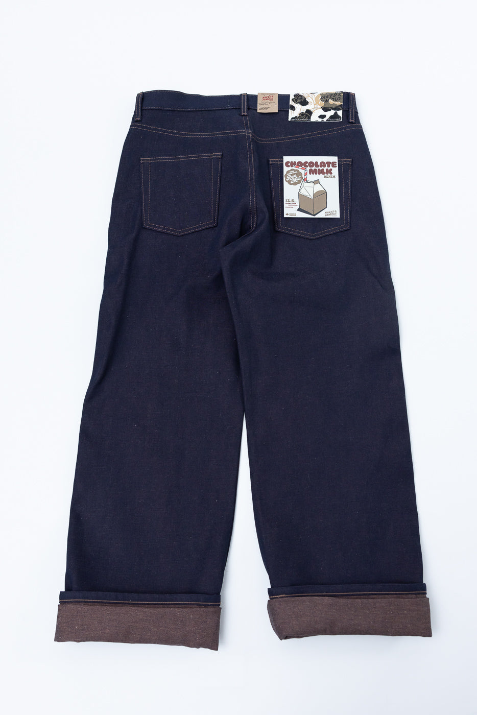 Flat lay of selvedge denim jeans, back side view, with two rear pockets, branded patch, visible tag, and brown cuff details.