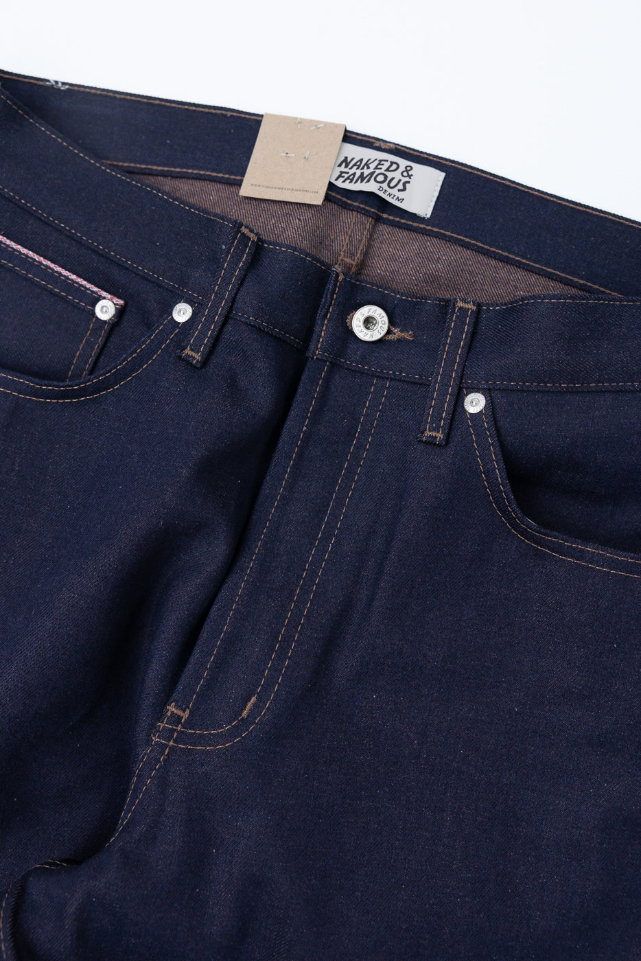 Close-up of the front waist and upper thigh of dark selvedge denim jeans, showing button, pockets, contrast stitching, and fabric texture.