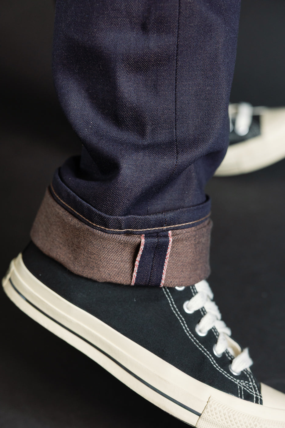 Close-up of male model's lower leg showing selvedge denim cuff and sneaker, side view, detailed stitching visible
