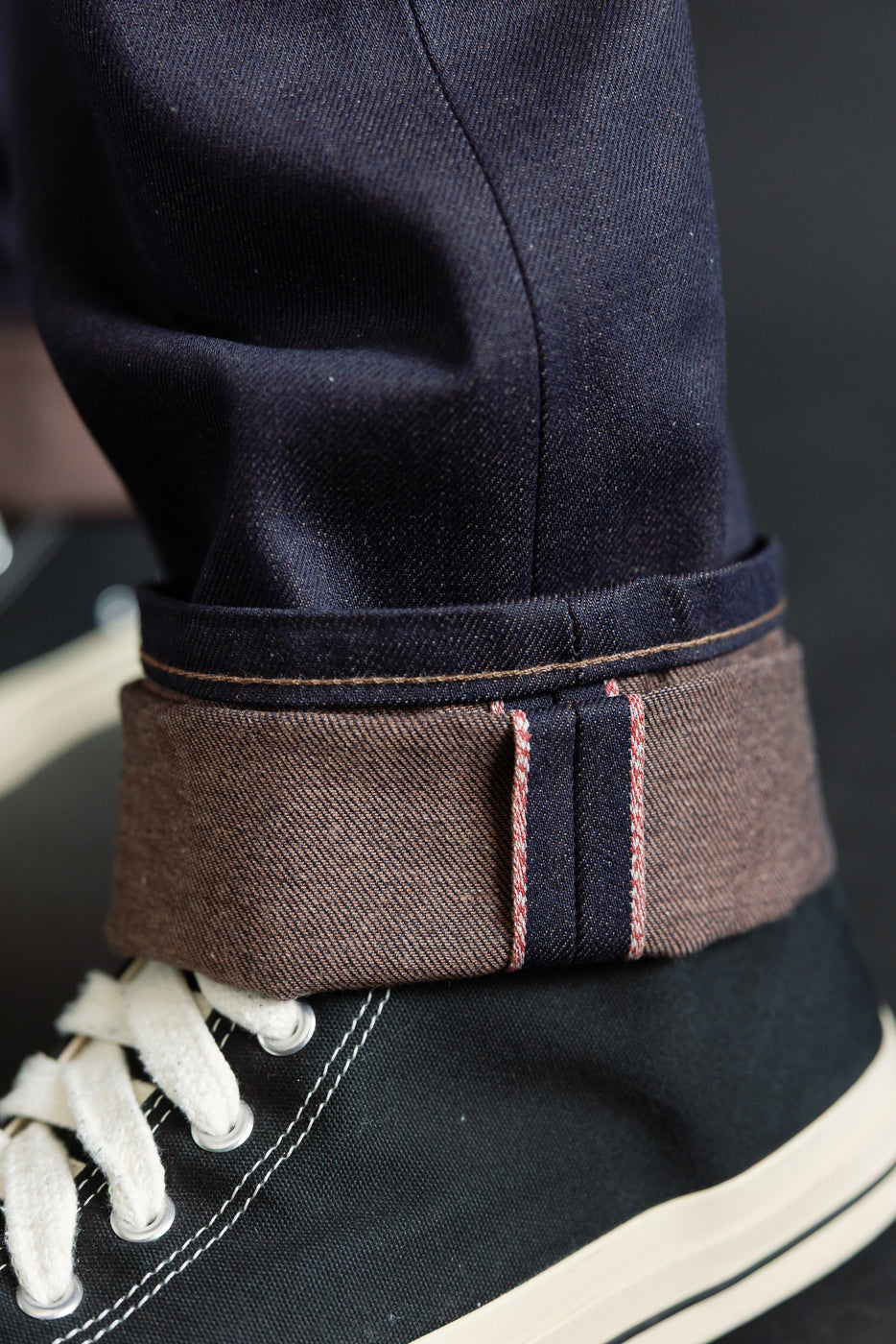 Close-up side view of men's selvedge jean cuff with visible chocolate brown weft and red line detail, model is 5'9
