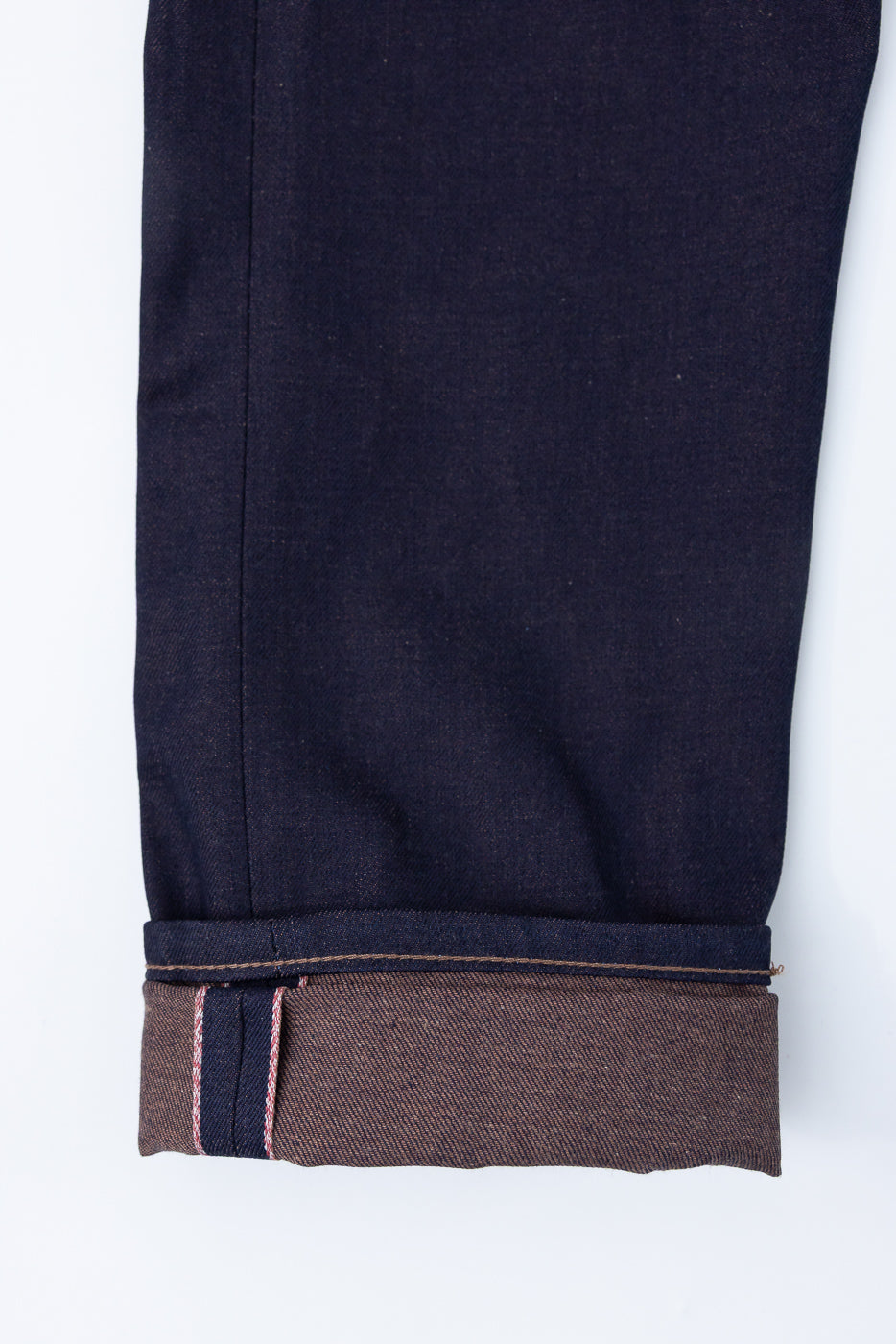 Close-up of selvedge denim pant leg cuff showing brown weft and red edge stitching, front view.