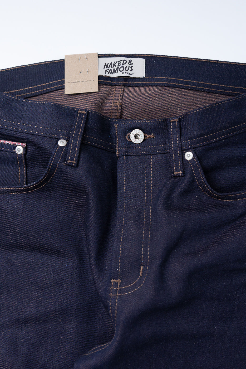 Close-up front view of men's dark selvedge denim jeans, showing button, stitching, and waistband tag.