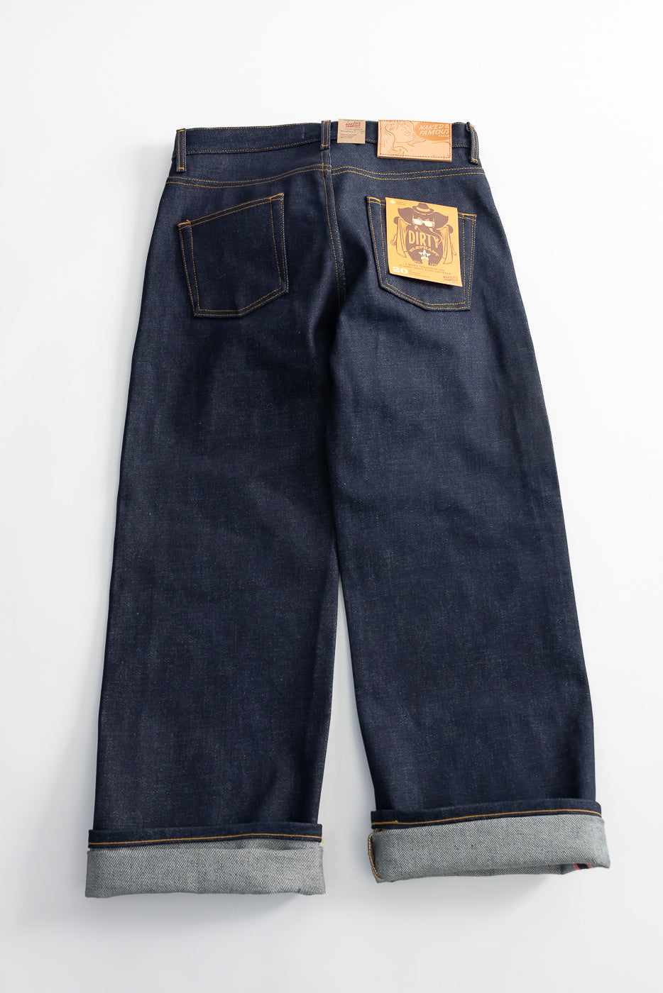 Flat lay of Naked & Famous Dirty Elephant 14oz selvedge jeans, back view, cuffs rolled, showing leather patch and rear pocket tag, from Dant contemporary casual shop.