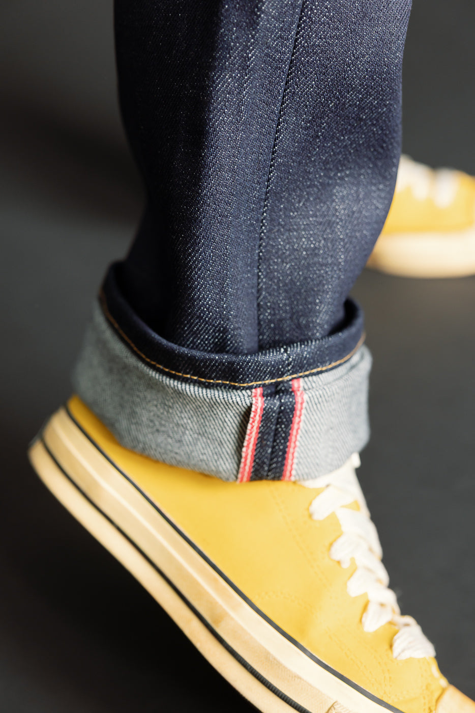 Close-up of male model cuffed Naked & Famous Dirty Elephant 14oz selvedge denim hem, left leg side view, paired with yellow sneaker; visible red selvedge line and textured indigo fabric.