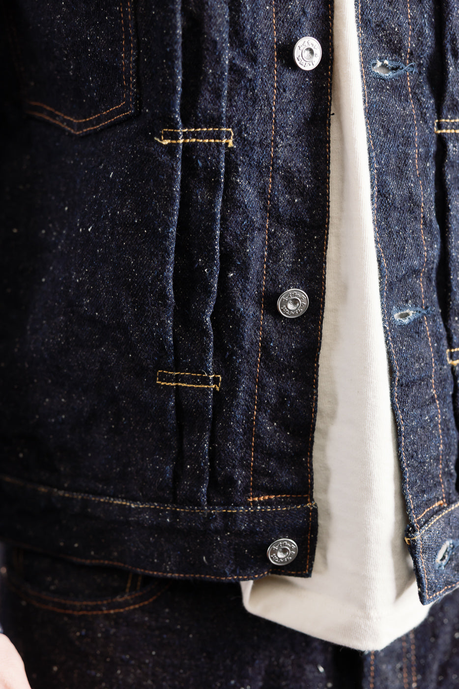 Close-up front view of Naked & Famous Genseki Selvedge denim jacket, showing metal buttons, textured dark indigo fabric, and yellow contrast stitching. Male model, 5'9