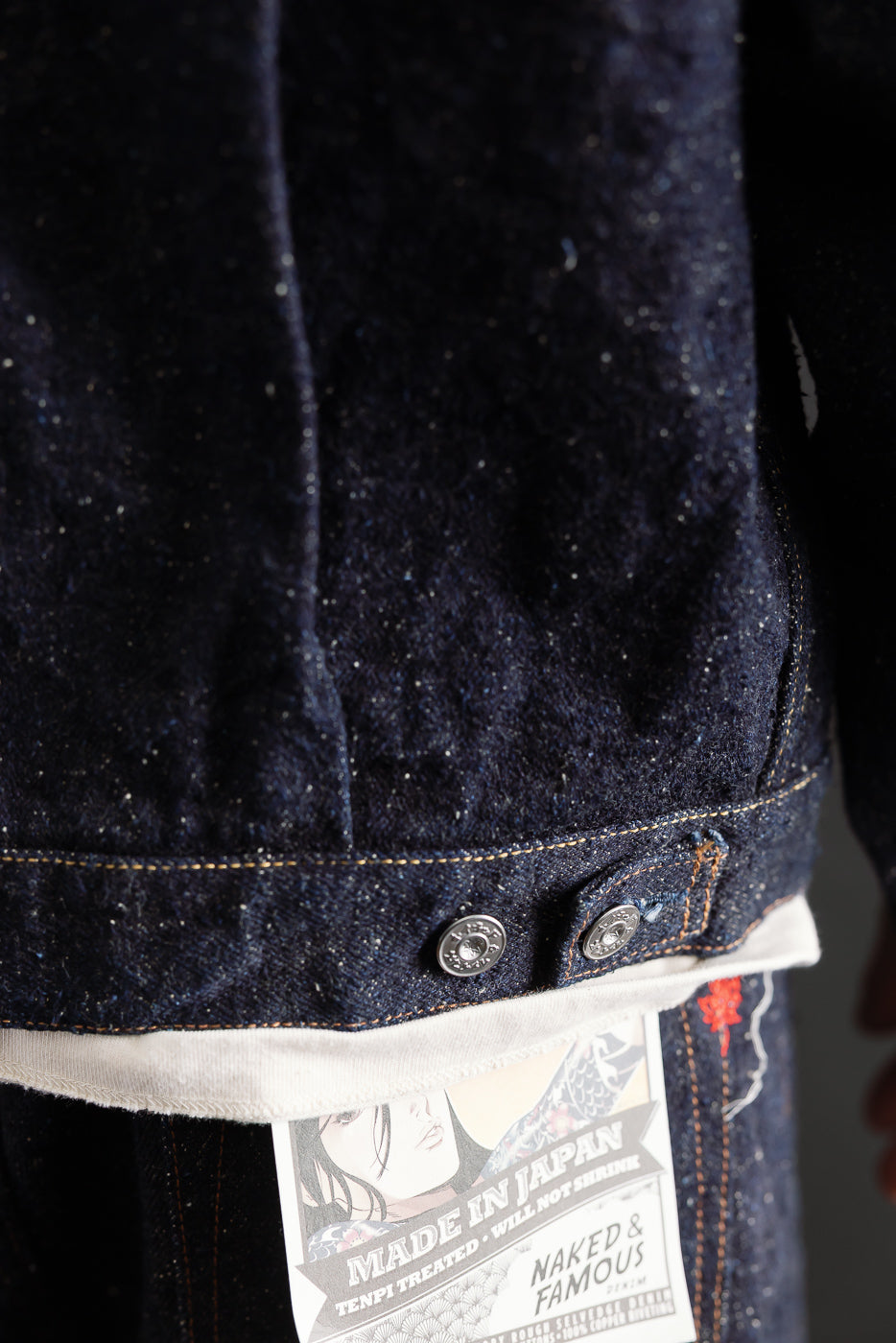 Close-up of Naked & Famous MIJ16 Genseki Selvedge heritage denim jacket back hem, showing button details, textured indigo fabric, and brand tag. Male model 5'9