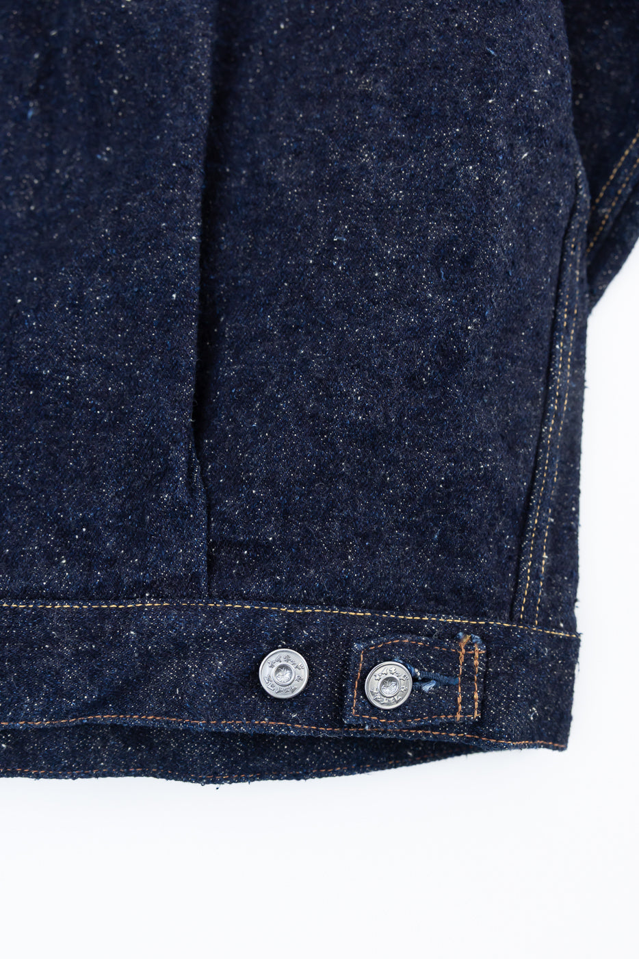 Close-up of back hem and waist buttons on men's Genseki Selvedge denim jacket, showing textured raw indigo fabric and contrast stitching.