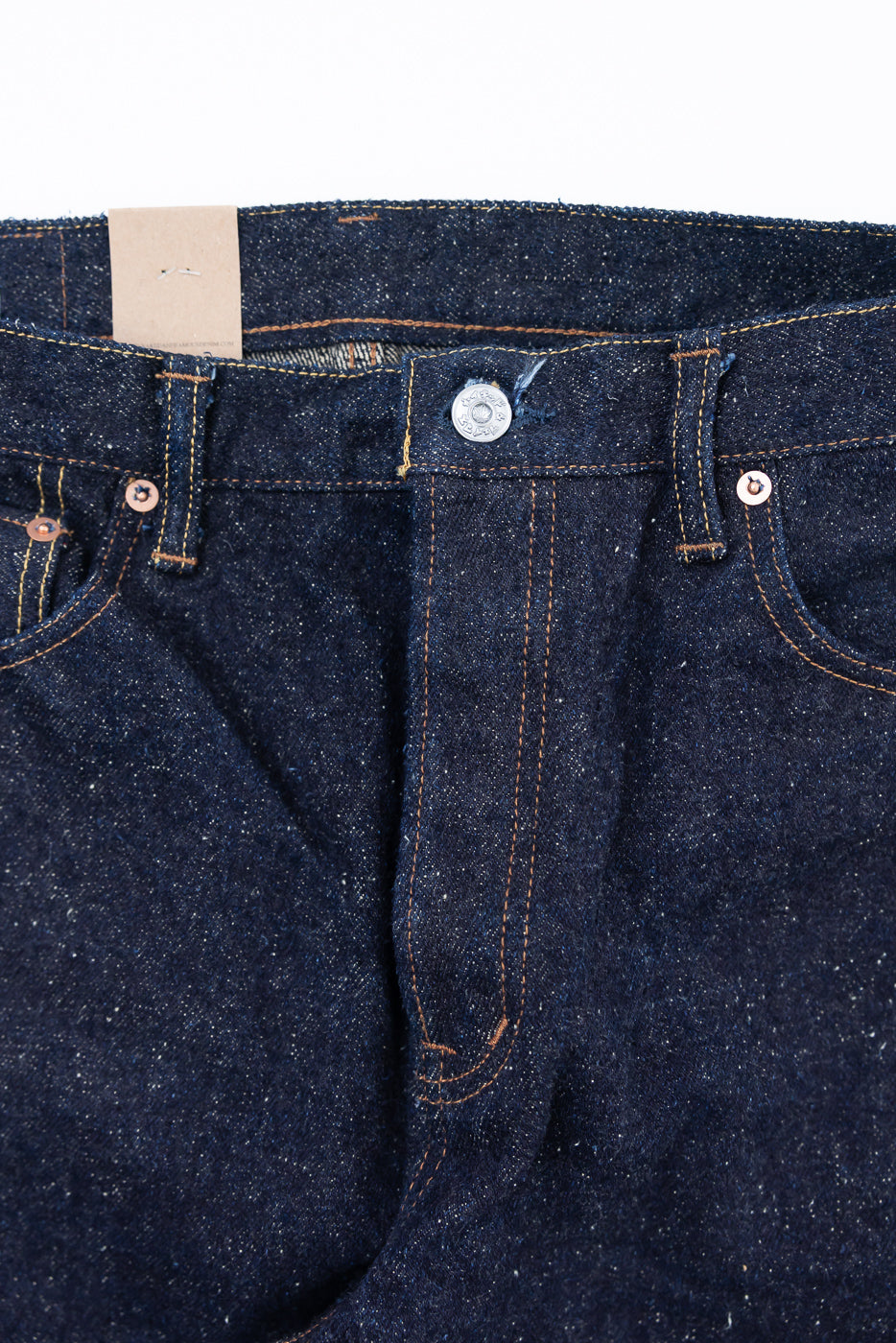 Close-up of front waist and fly of Naked & Famous selvedge denim jeans, showing button, rivets, and blue textured fabric.