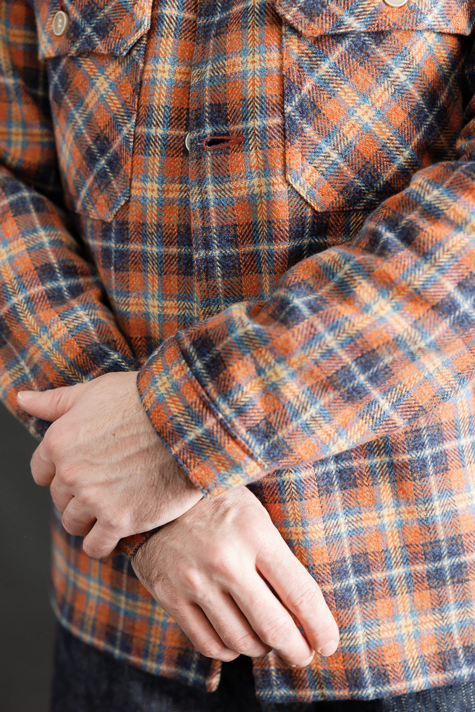 Close-up of male model's torso in orange plaid work shirt, showcasing chest pockets and cuff details.