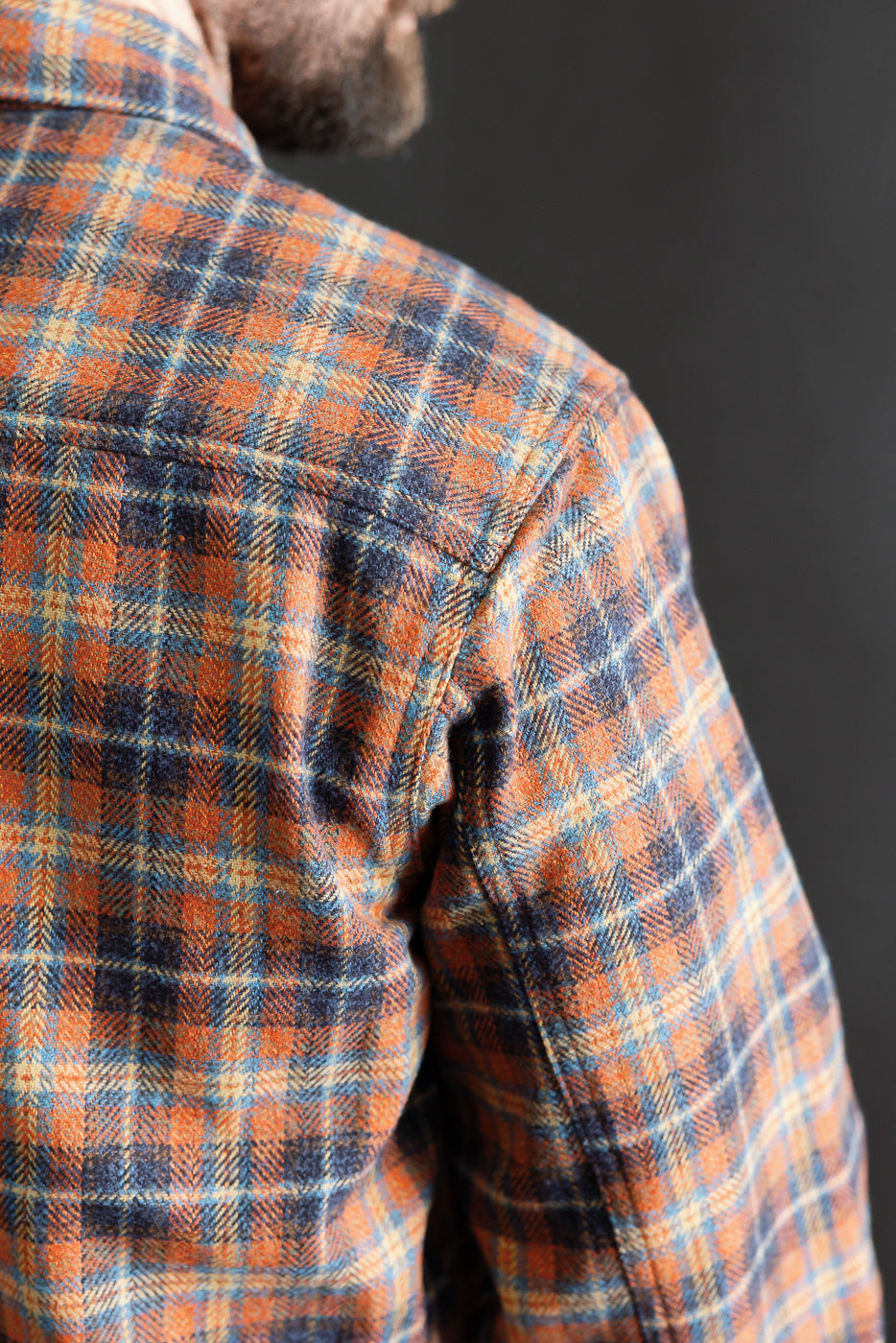 Close-up of male model's upper back and shoulder wearing Naked & Famous plaid work shirt, highlighting orange, blue, and tan triple twist yarn fabric texture, seen from the back.