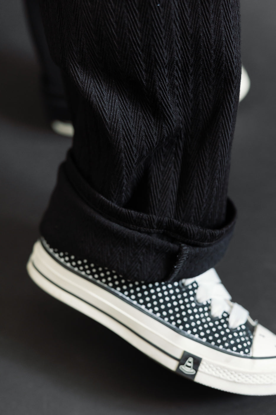 Close-up of the cuffed hem of black herringbone texture trousers and polka dot sneakers.