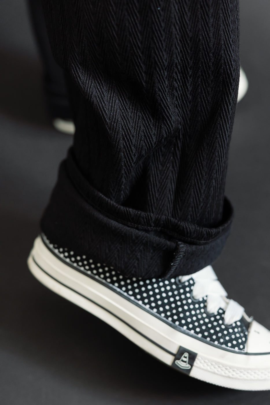 Close-up of the bottom hem and cuff of black herringbone patterned trousers with polka dot sneakers.