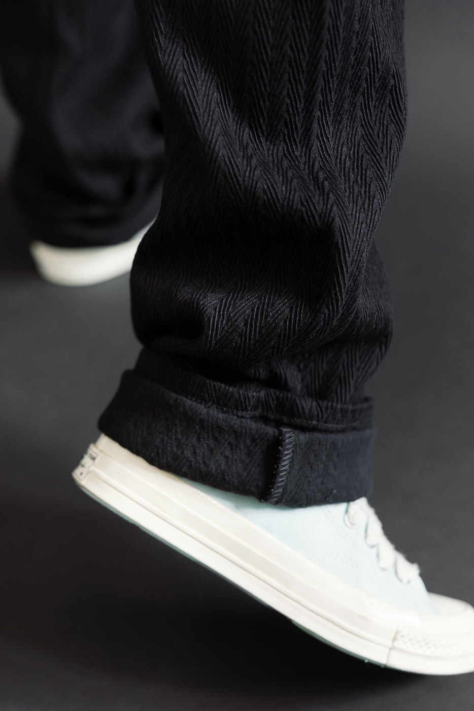 Close-up of the bottom hem and cuffed leg of black textured trousers paired with white sneakers.