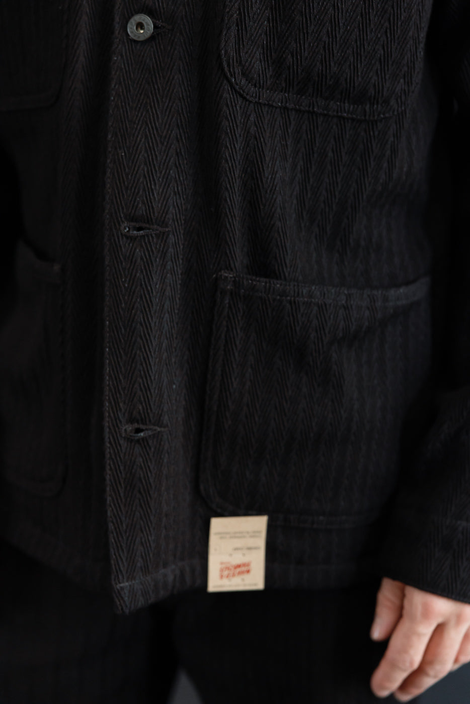 Close-up, front view of black herringbone fabric texture, large patch pocket, and bottom hem tag.