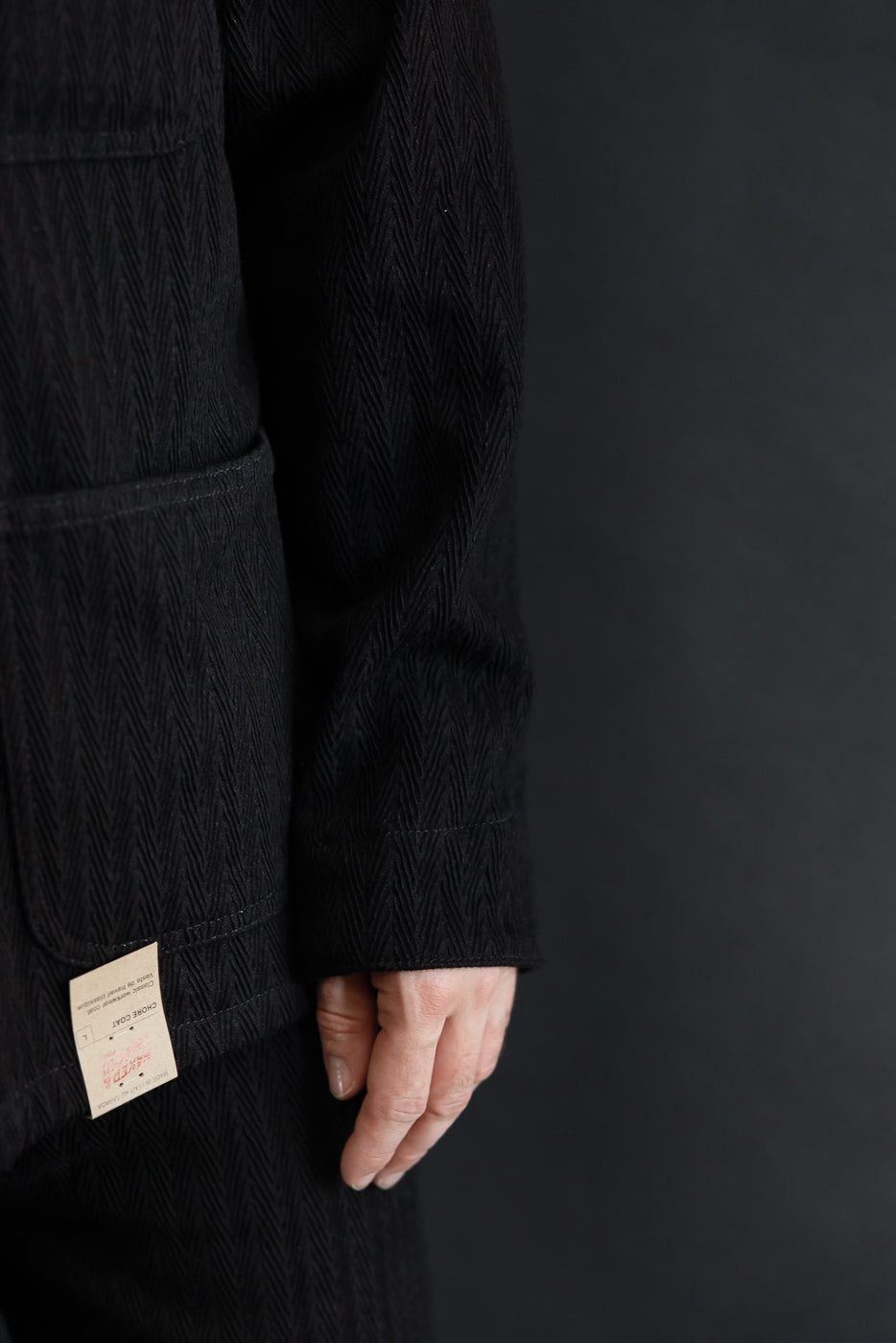 Close-up side view of a black herringbone fabric chore coat sleeve and hem detail.
