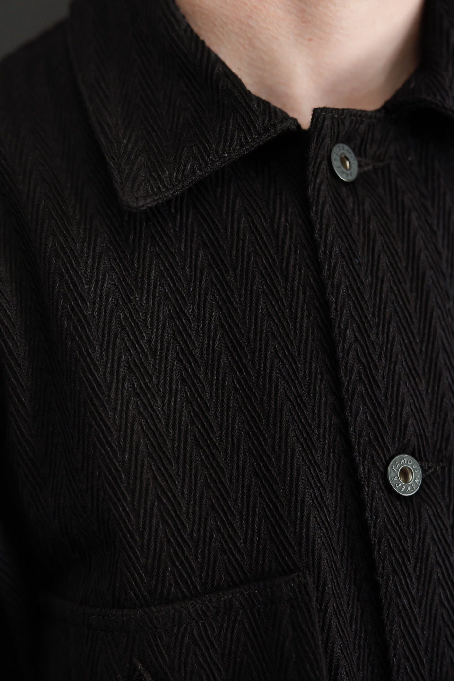 Close-up front view of black herringbone textured fabric with collar and metal button details.