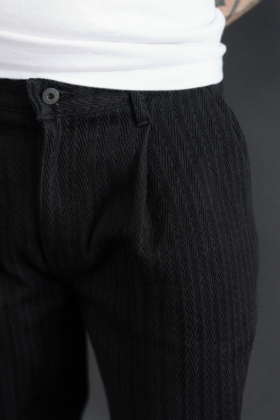 Close-up front view of black herringbone pattern trousers on a male model, showing waistband and button.
