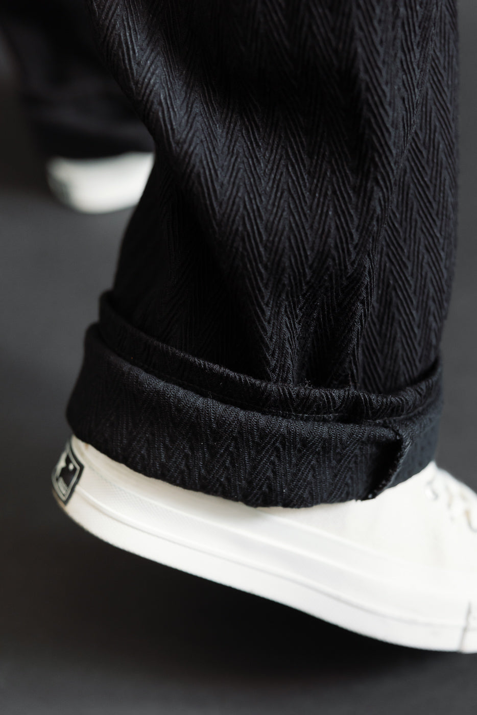 Close-up of male model's left pant leg hem with herringbone texture and white sneakers.