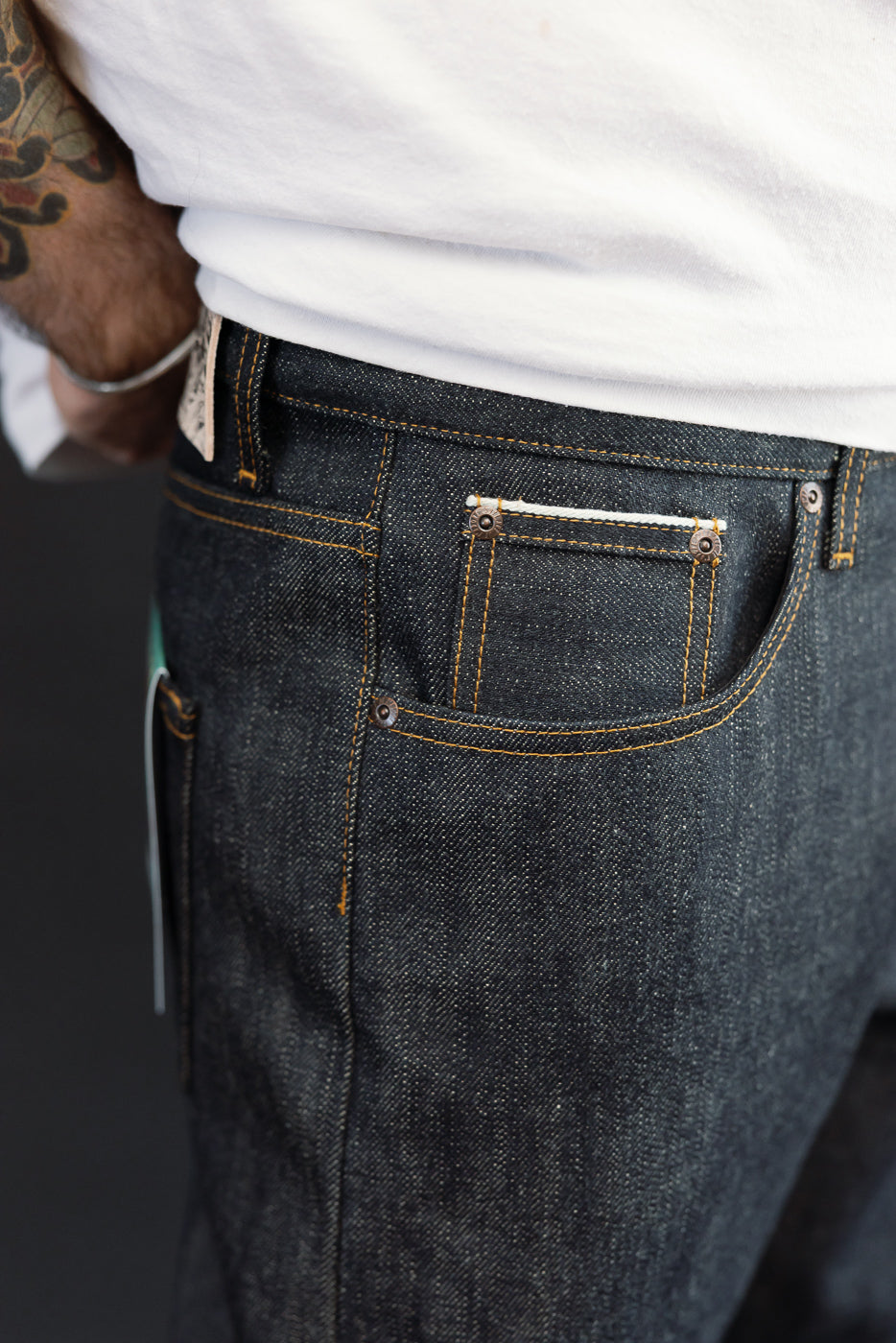 Close-up of male model's right side showing dark selvedge denim jeans with orange stitching, back pocket, and metal rivets