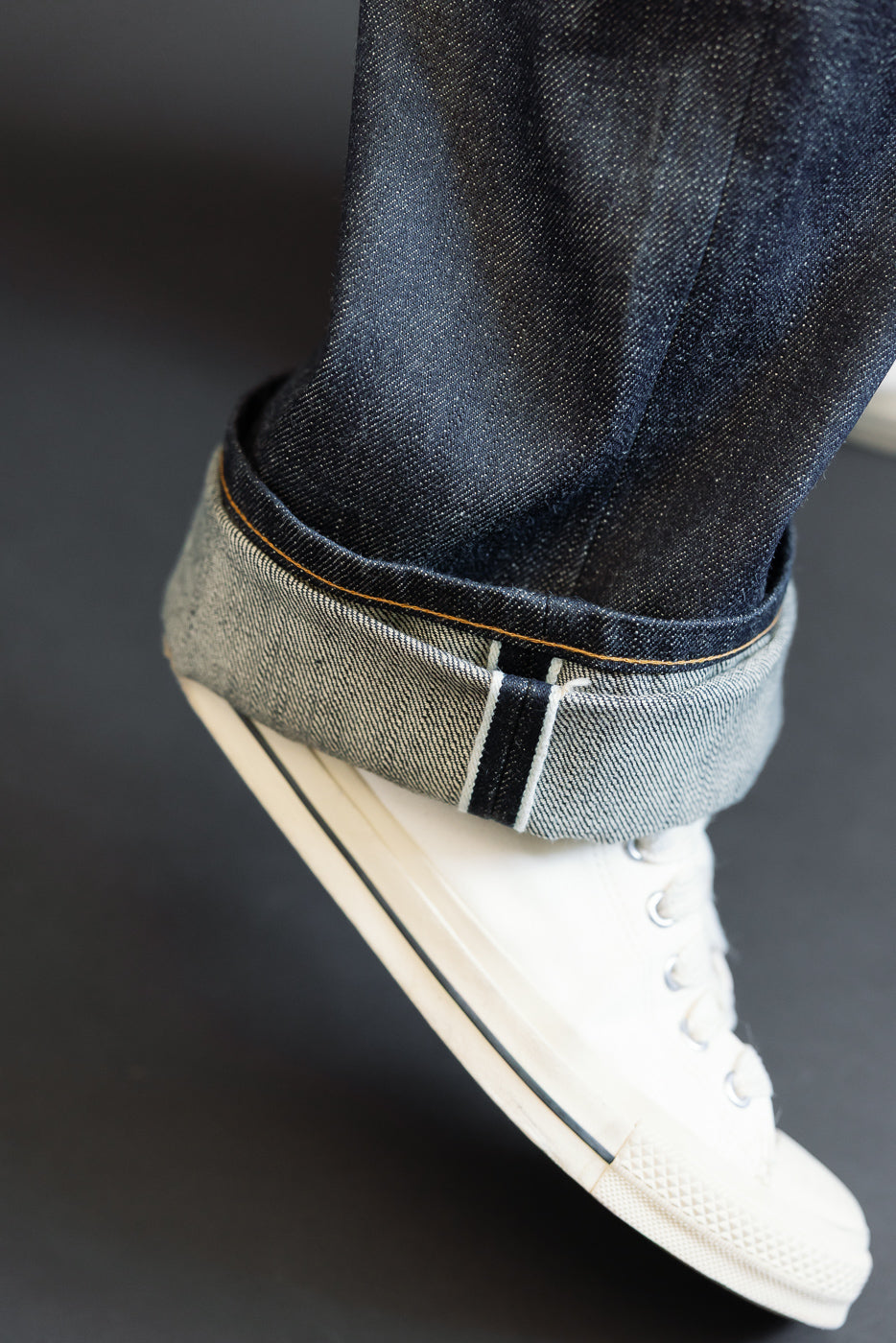 Close-up side view of male model's cuffed selvedge denim hem with orange stitching, paired with white sneakers.
