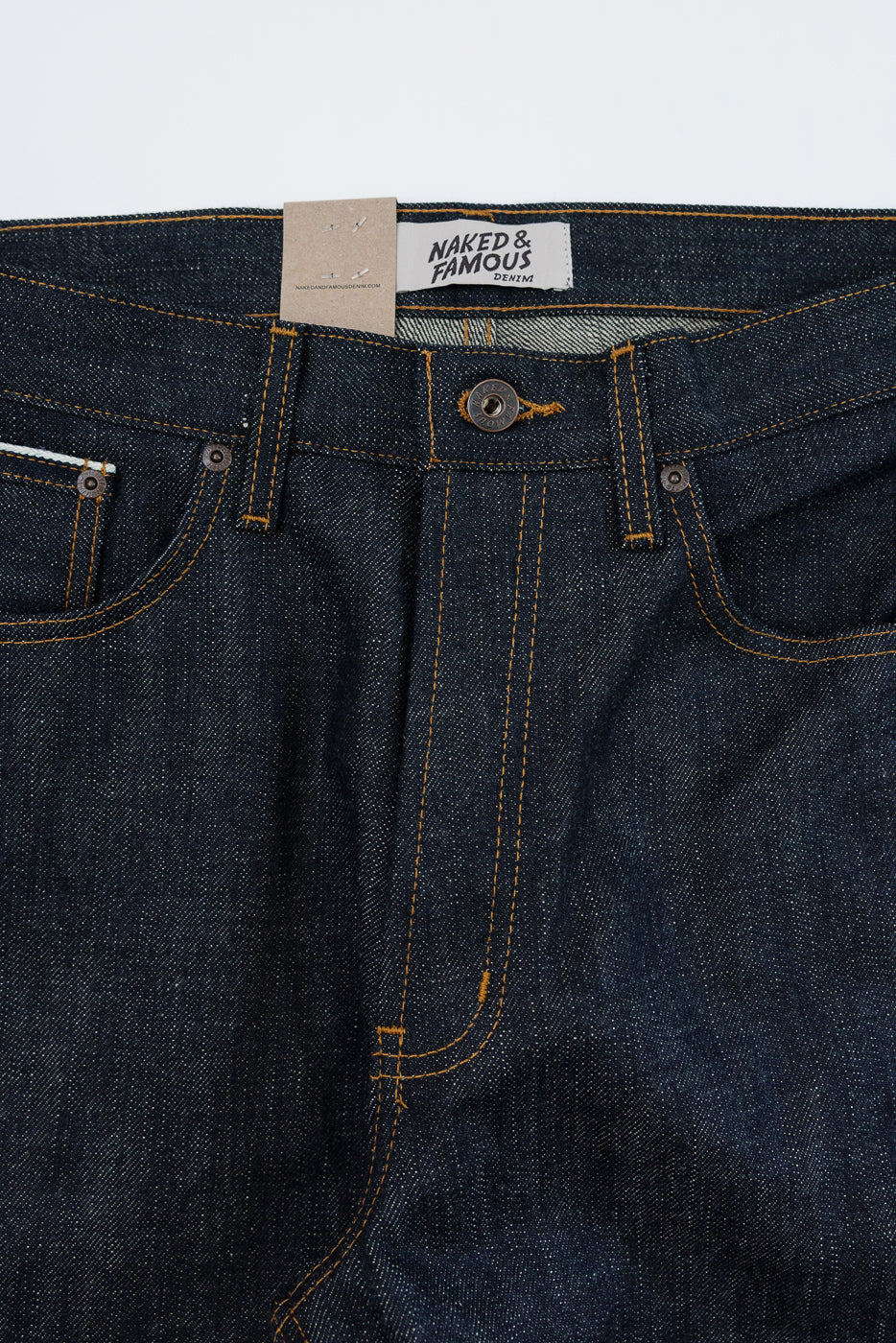 Close-up front view of dark selvedge denim jeans with orange stitching, button fly, and Naked & Famous tag.