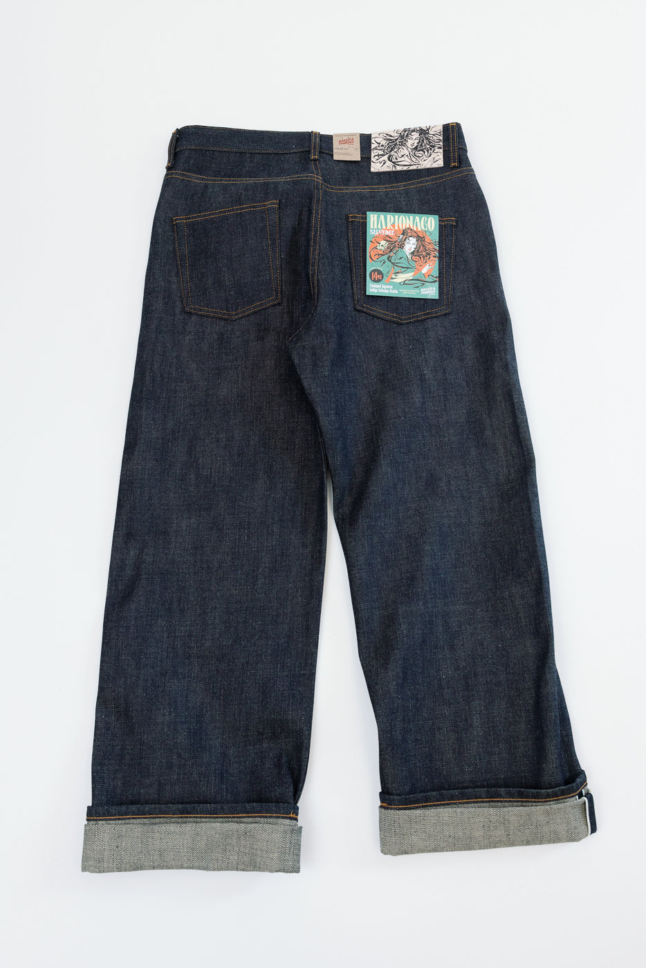 Back view flat lay of dark selvedge denim jeans with rolled hems, branded back pocket patch, and waist label