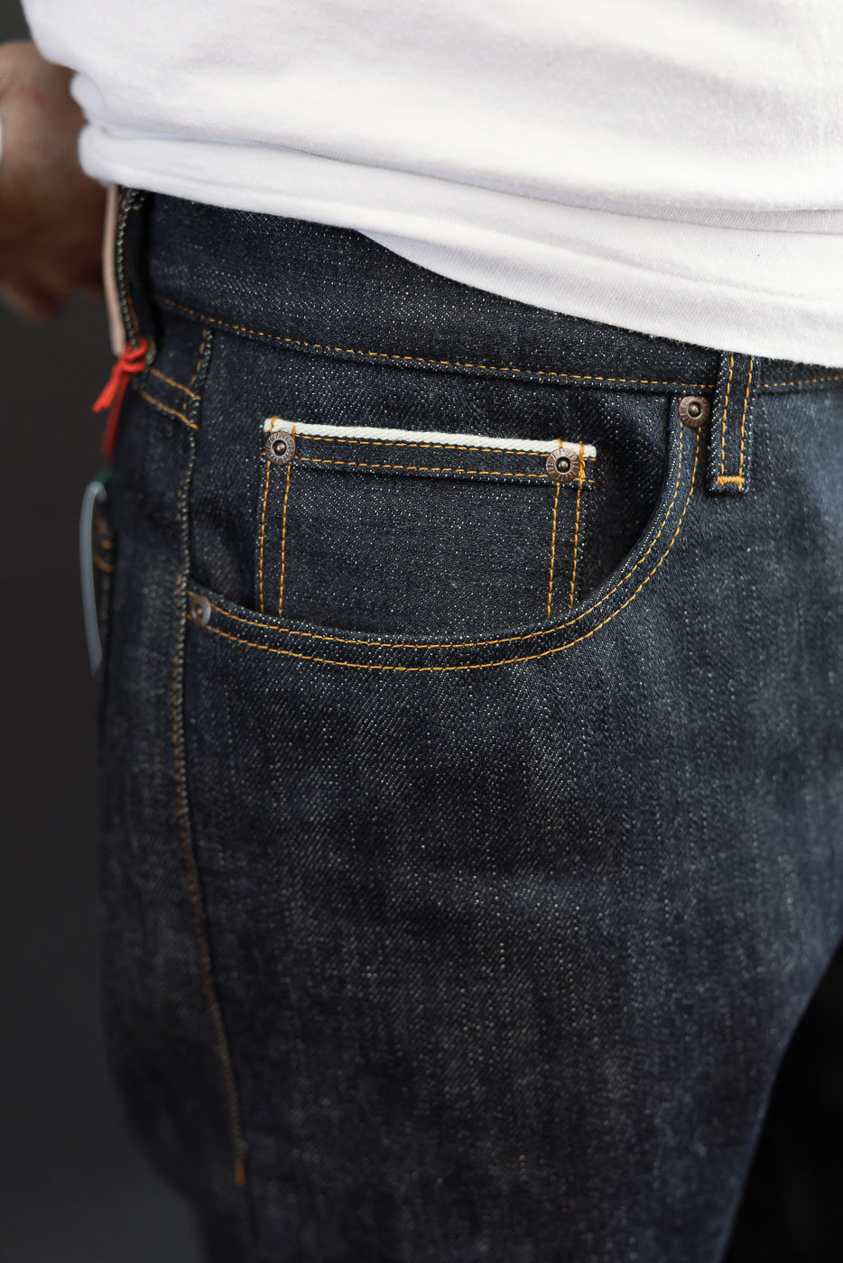 Close-up back view of dark selvedge denim jeans on male model, showing rear pocket with contrast stitching and copper rivets