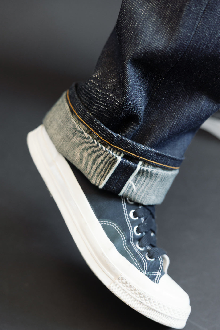Close-up side view of male model's cuffed selvedge denim hem with orange stitching, paired with dark canvas sneakers