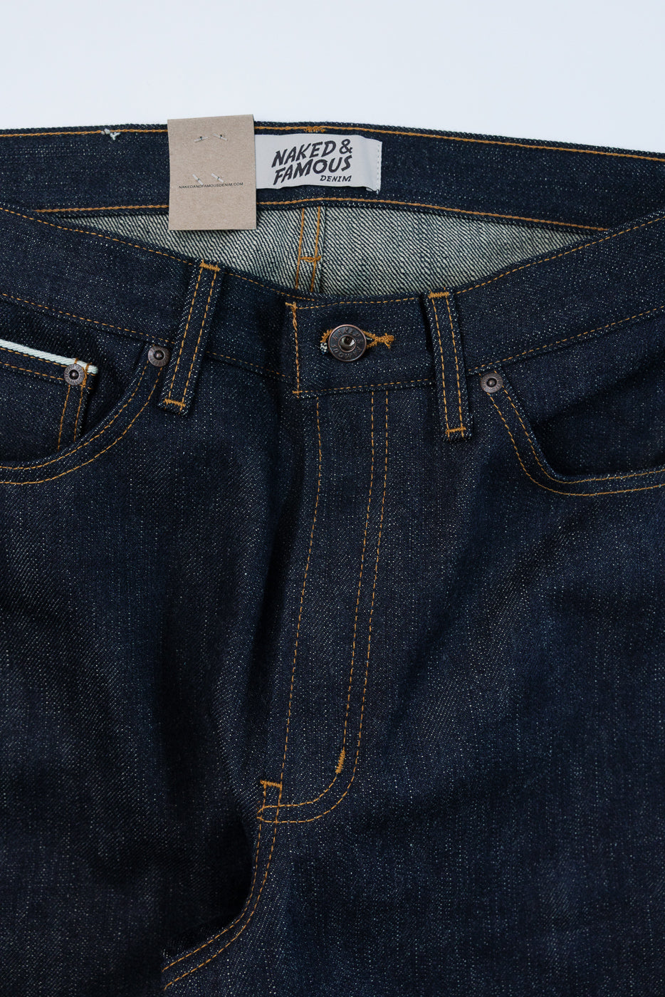 Close-up front view of dark selvedge denim jeans showing Naked & Famous tag, orange stitching, button fly, and pocket details