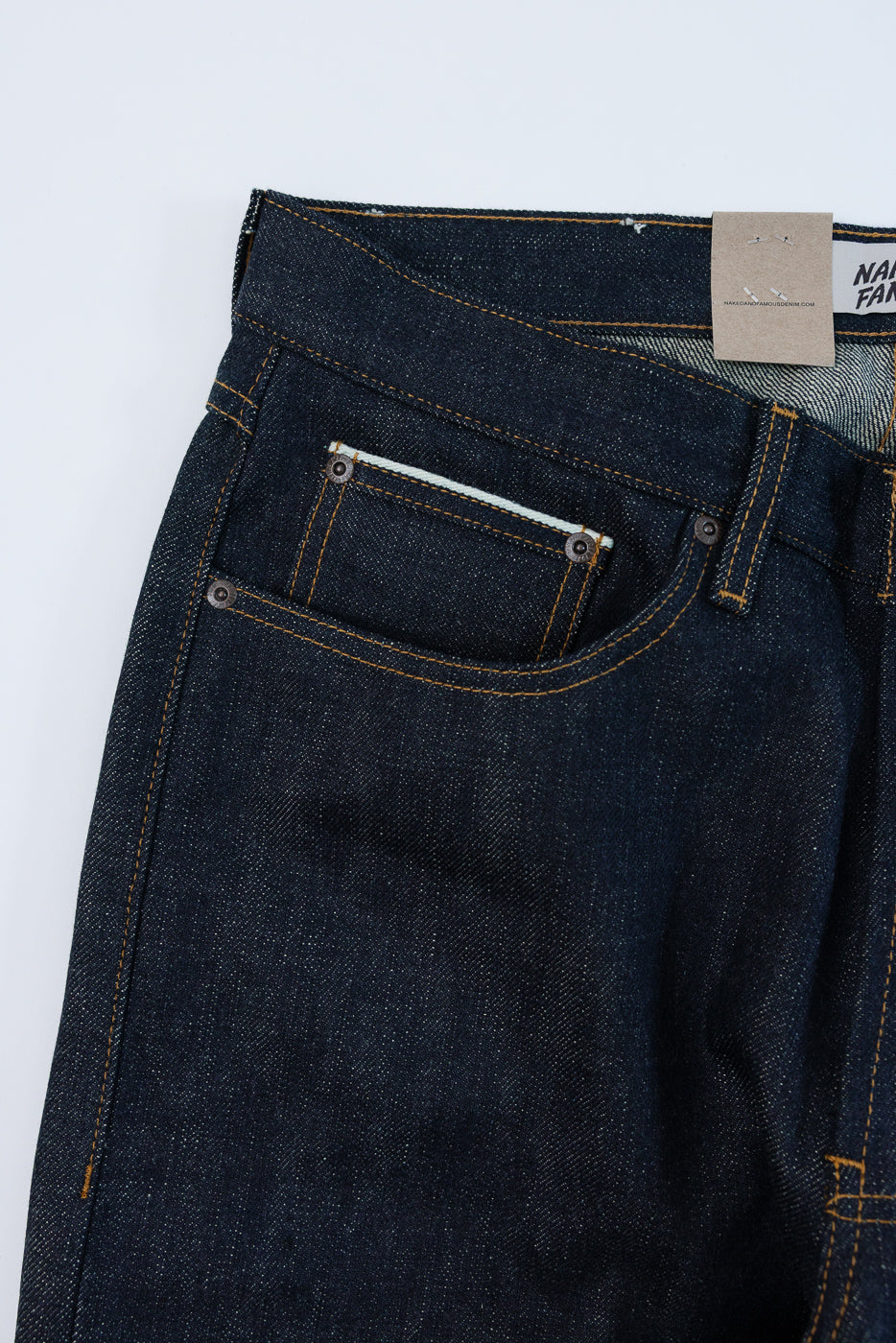 Close-up front view of dark selvedge denim jeans, showcasing front pocket, orange stitching, and button details.