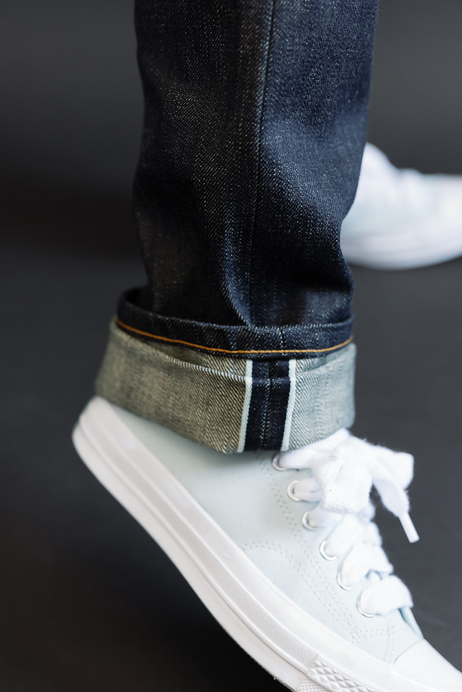 Close-up of male model's cuffed selvedge denim jeans showing raw hem, orange stitching, and contrast interior, paired with white sneakers