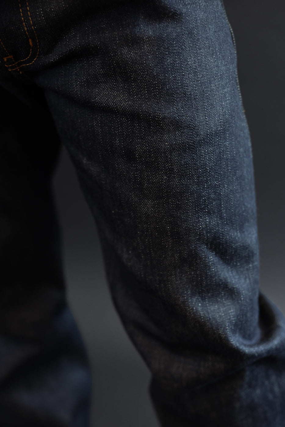 Close-up side view of dark selvedge denim jeans on male model, showcasing fabric texture and orange stitching detail
