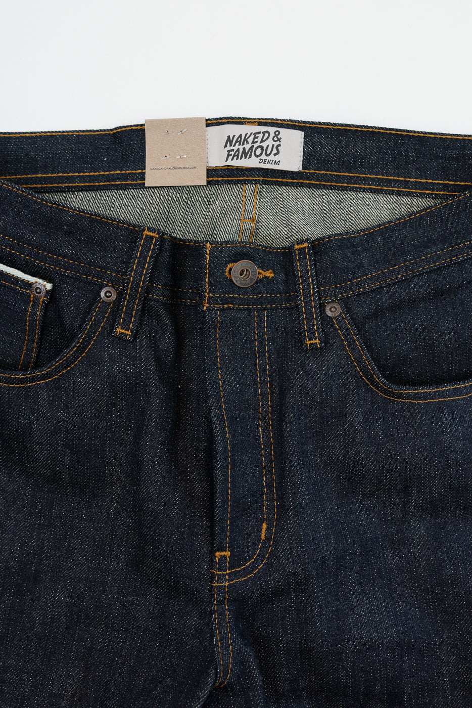 Close-up front view of dark selvedge denim jeans showing waistband, button fly, orange stitching, and Naked & Famous tag.