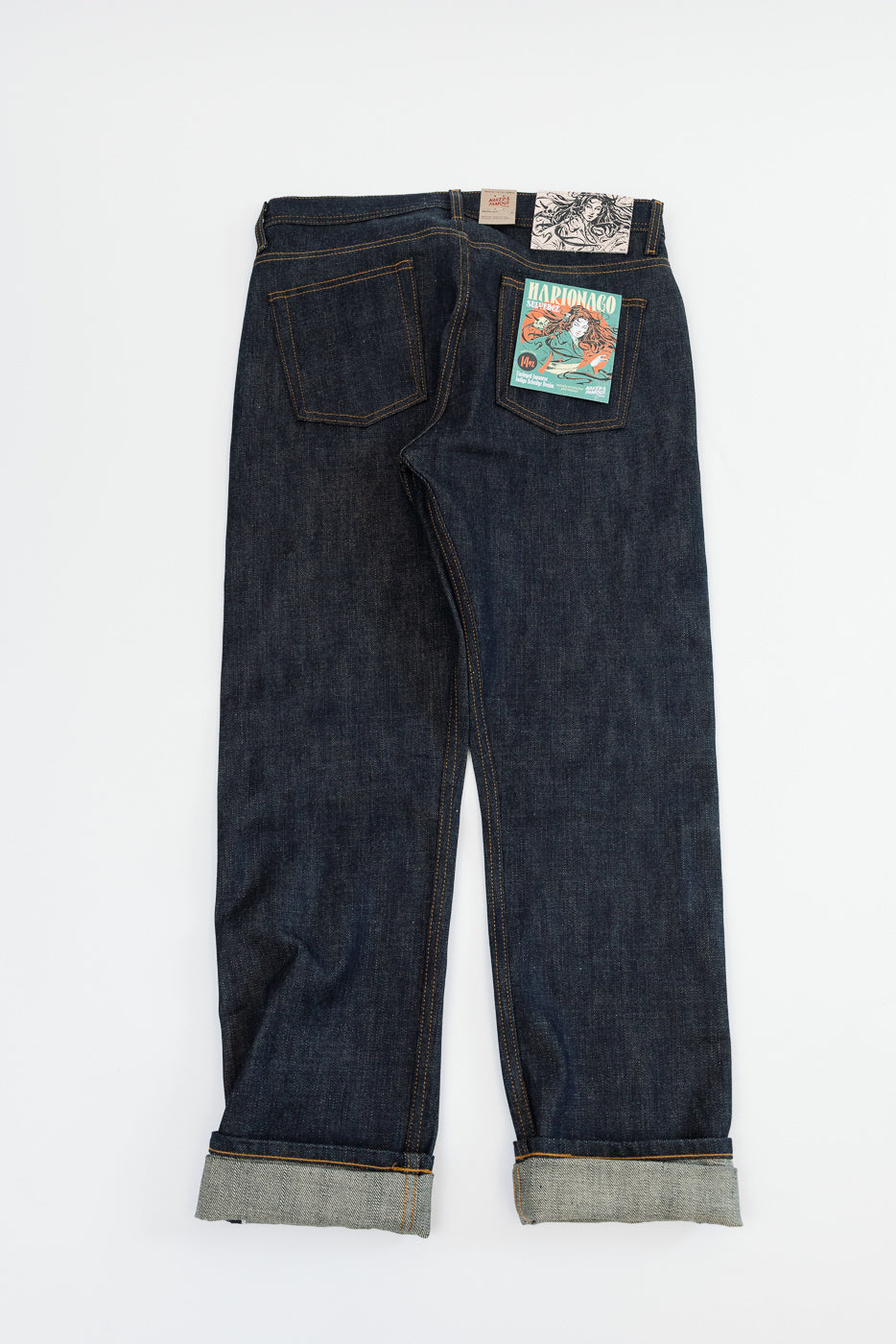Back view flat lay of dark selvedge denim jeans with cuffed hems, orange stitching, and graphic back pocket patch.