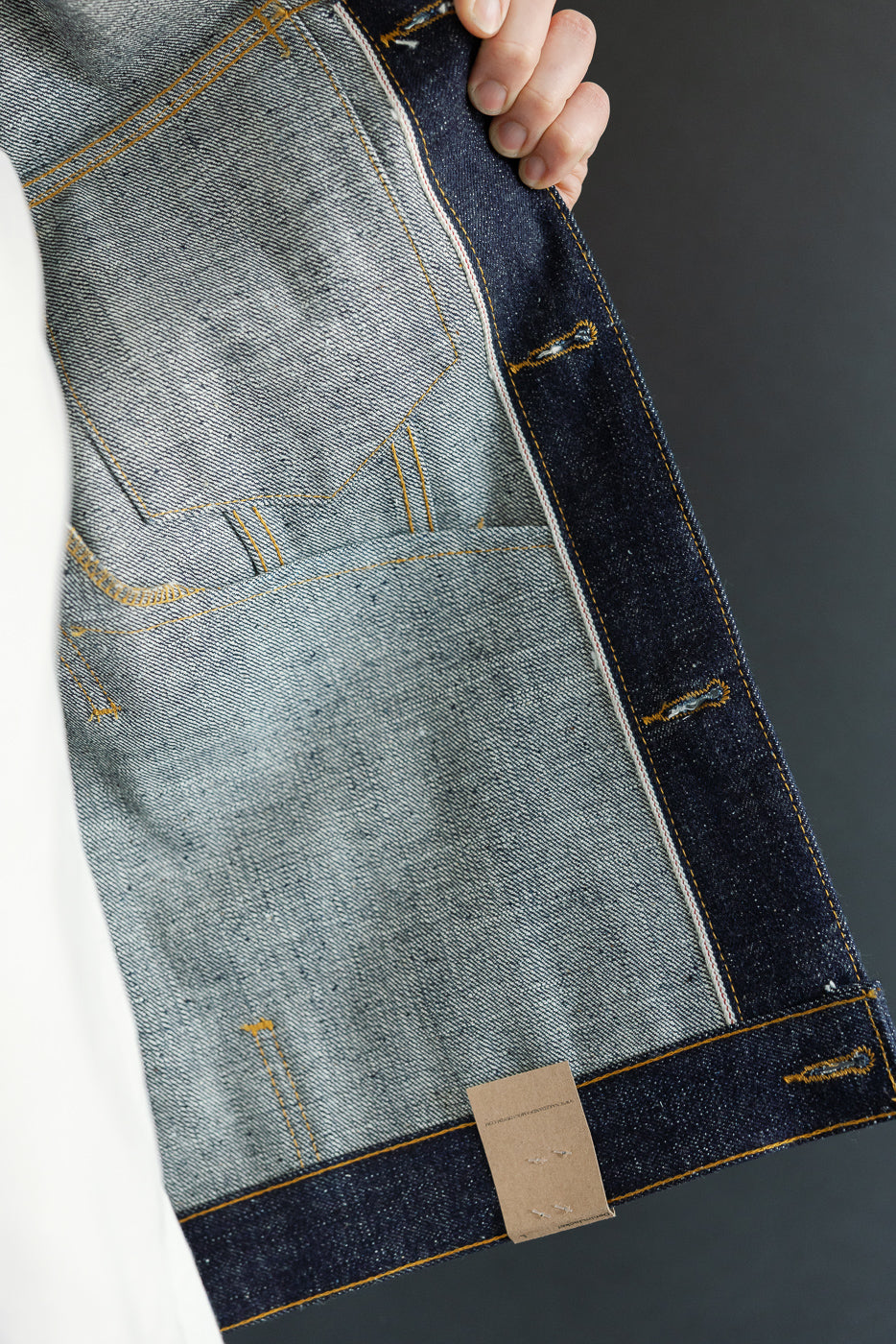Close-up of male model's denim jacket interior, showcasing selvedge fabric, yellow stitching, and tag details.