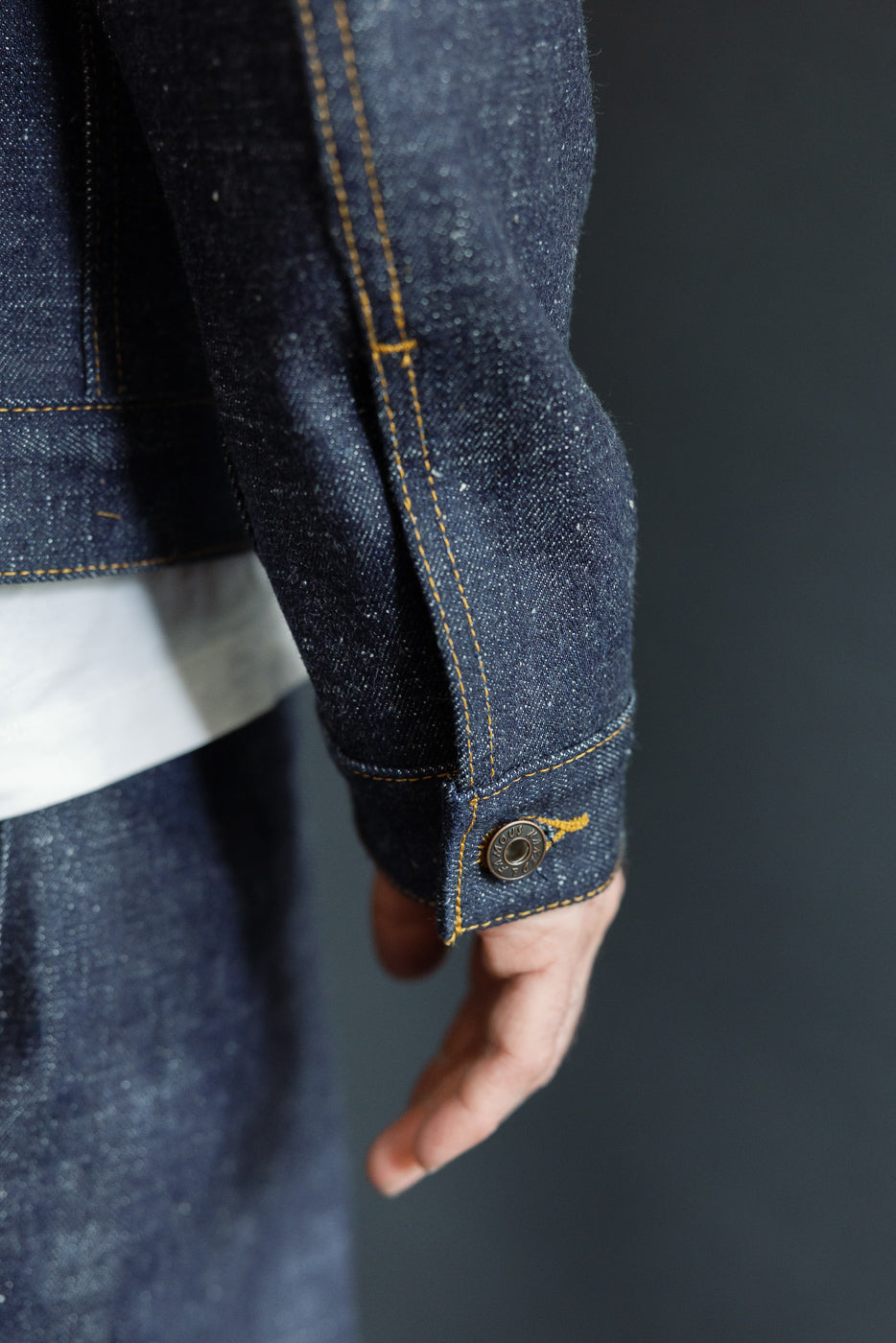 Close-up side view of male model's denim jacket sleeve, showing button cuff, gold stitching, and raw indigo fabric texture.