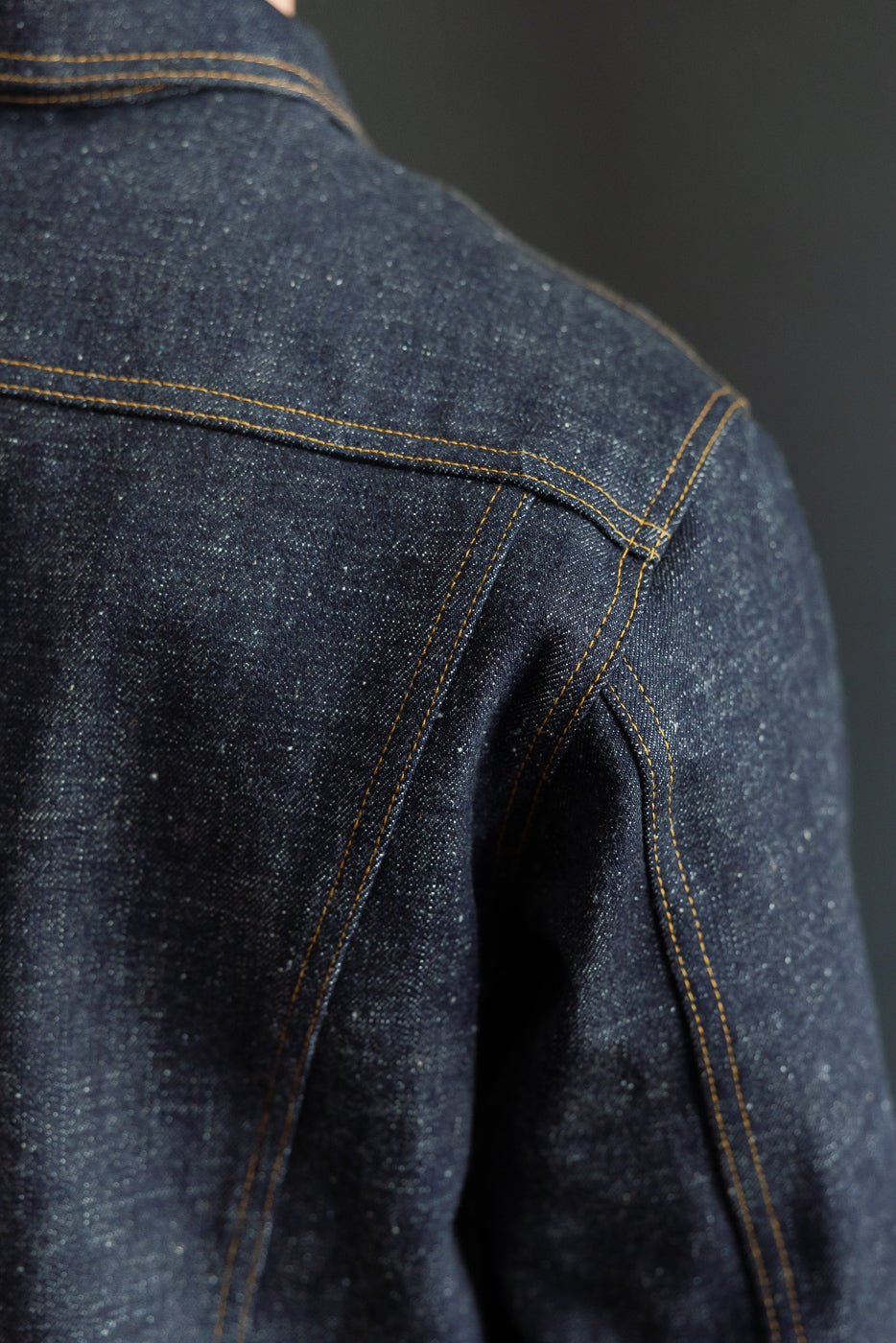 Close-up of male model's shoulder, showcasing dark denim jacket with orange stitching and fabric texture.