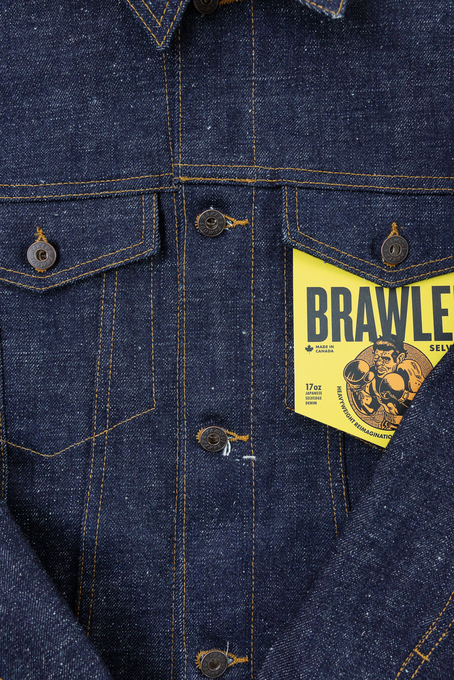 Close-up front of men's 17oz Japanese selvedge denim jacket, showing chest pockets, brass buttons, and yellow BRAWLE tag.