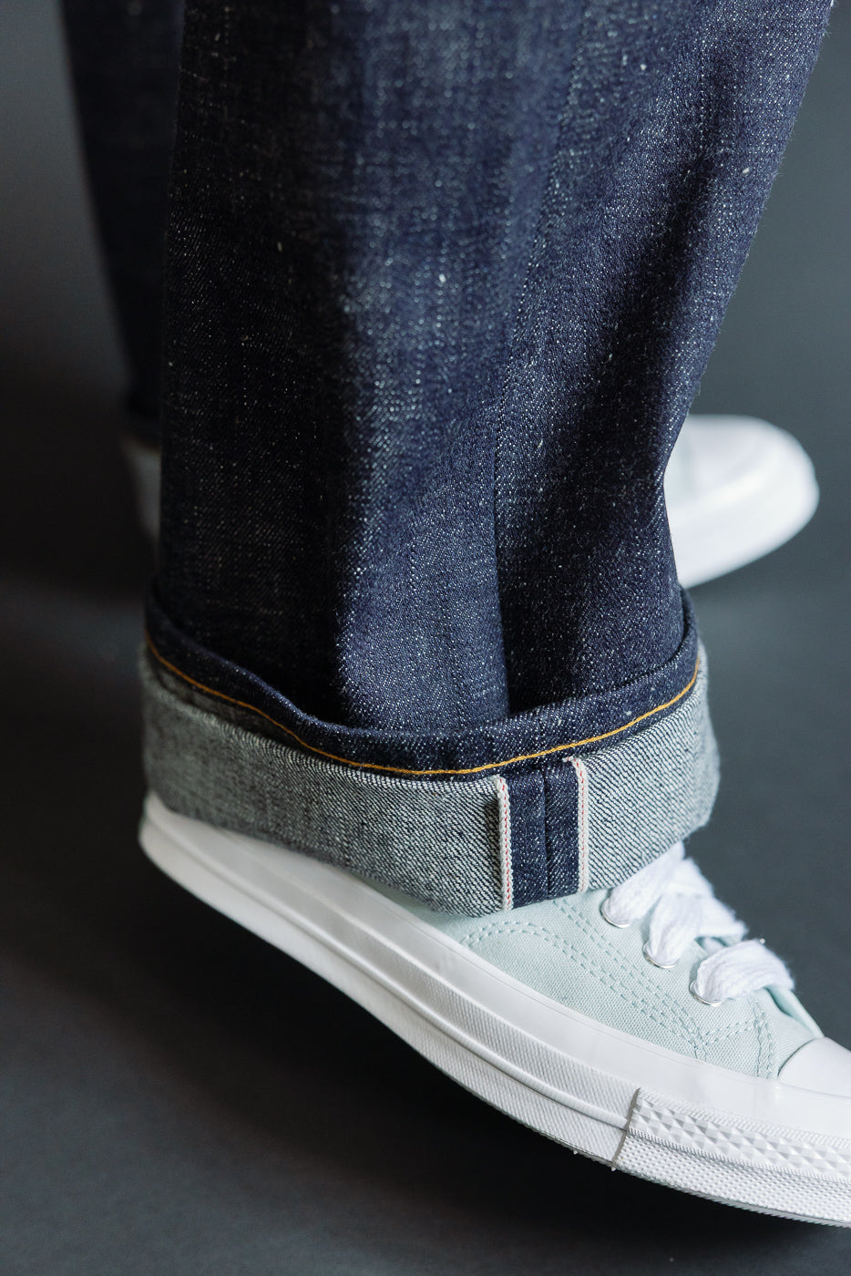Close-up of male model's cuffed selvedge denim jeans, showing raw hem and contrast stitching with light sneakers
