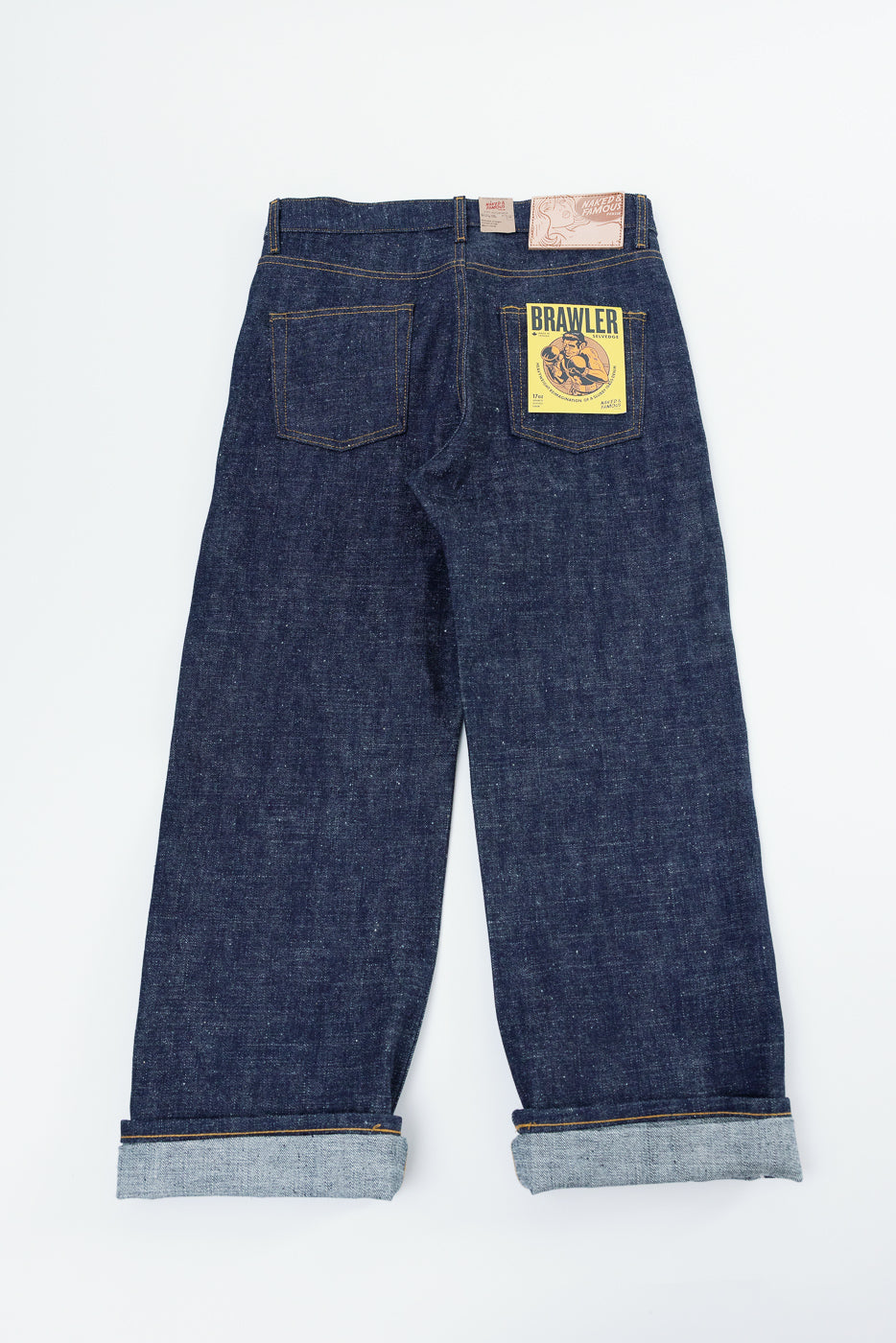 Back view flat lay of dark selvedge denim jeans with 'BRAWLER' patch, cuffed hems, and yellow stitching