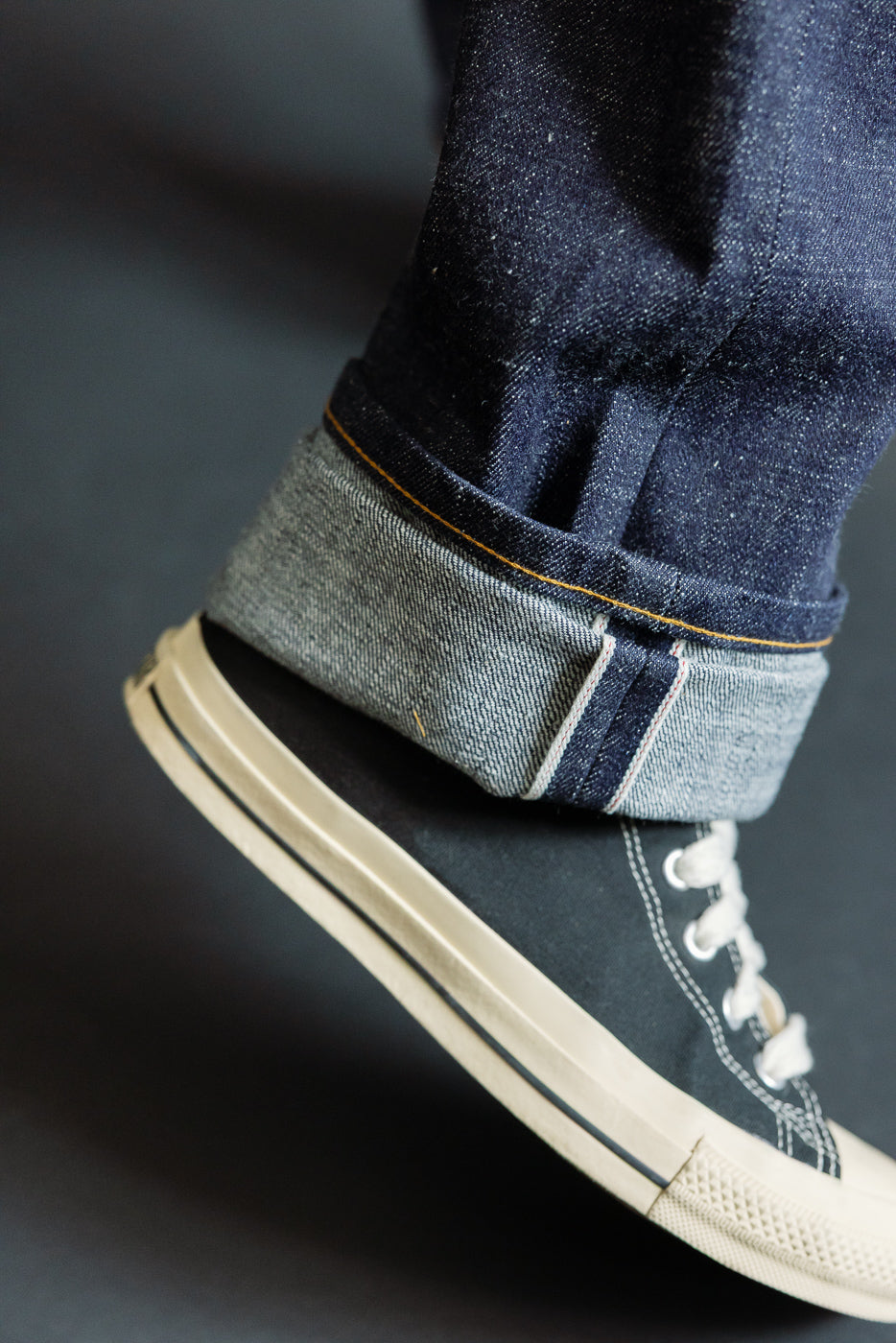 Close-up of male model's cuffed selvedge denim hem with orange stitching, paired with black canvas sneakers