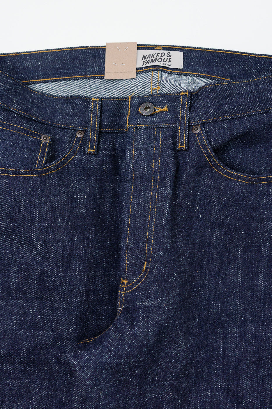 Close-up front of raw selvedge denim jeans, showing button fly, yellow stitching, and Naked & Famous tag.