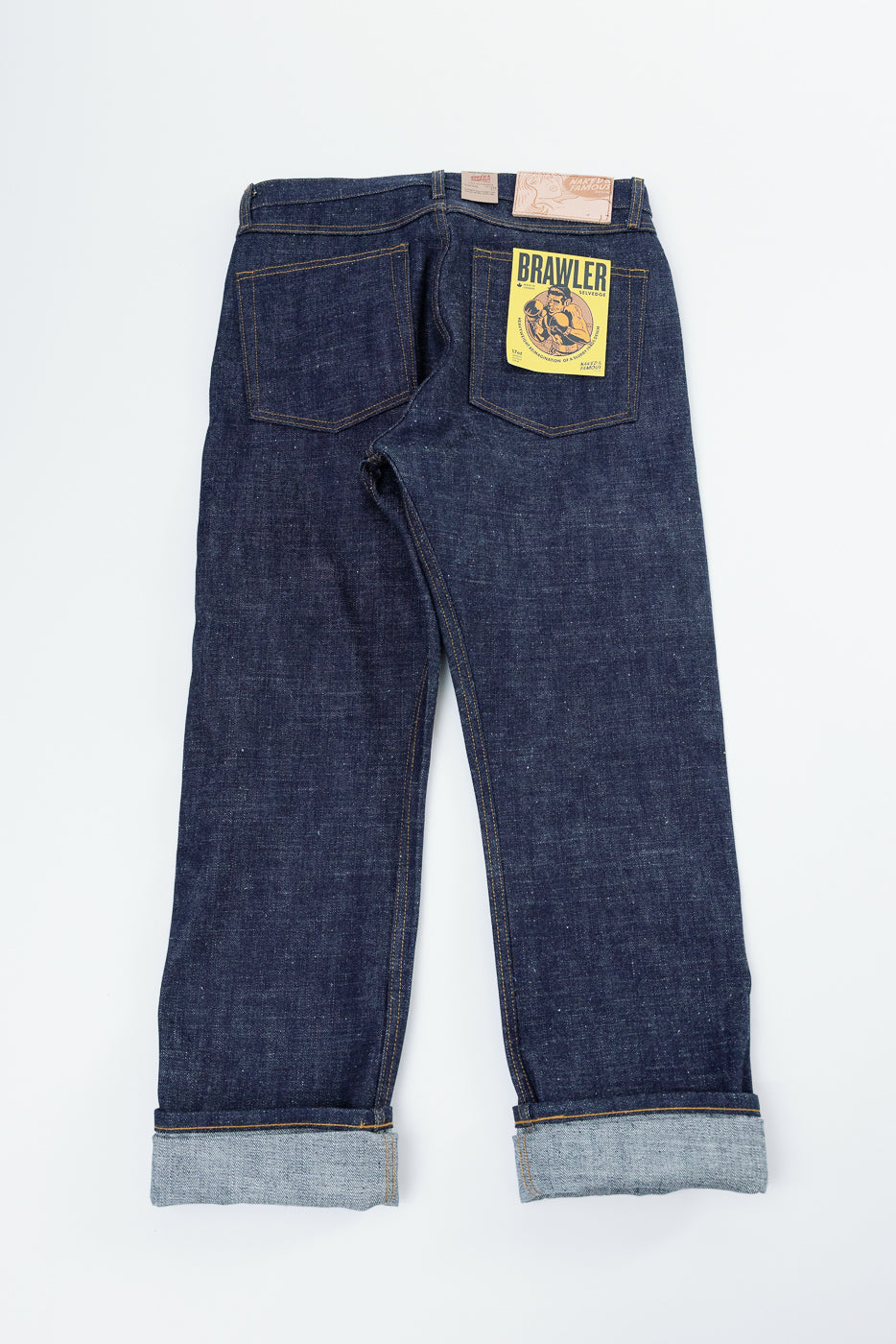 Back view flat lay of dark selvedge denim jeans with yellow Brawler patch, cuffed hems, and contrast stitching
