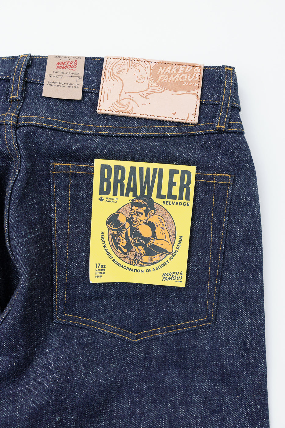 Close-up back view of Naked & Famous Brawler selvedge jeans, showing leather patch, care tag, and yellow pocket label on dark denim