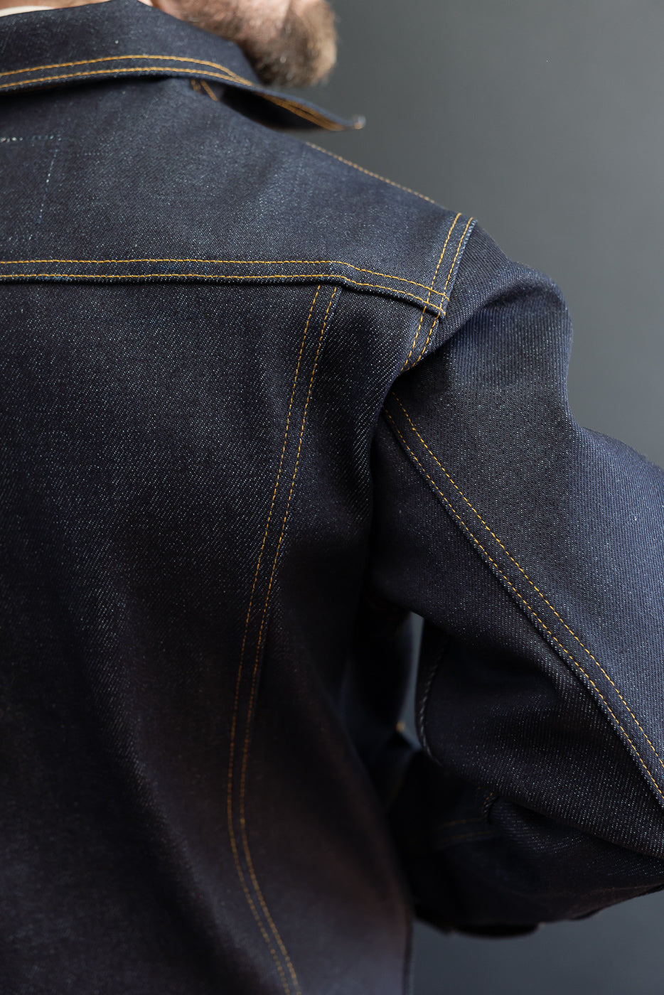 Close-up back view of a male model wearing a dark indigo selvedge denim jacket showing shoulder and sleeve stitching.
