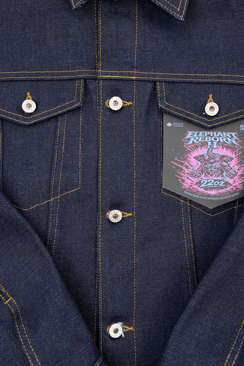 Close-up front view of a dark indigo selvedge denim jacket showing button details and a pink graphic tag.
