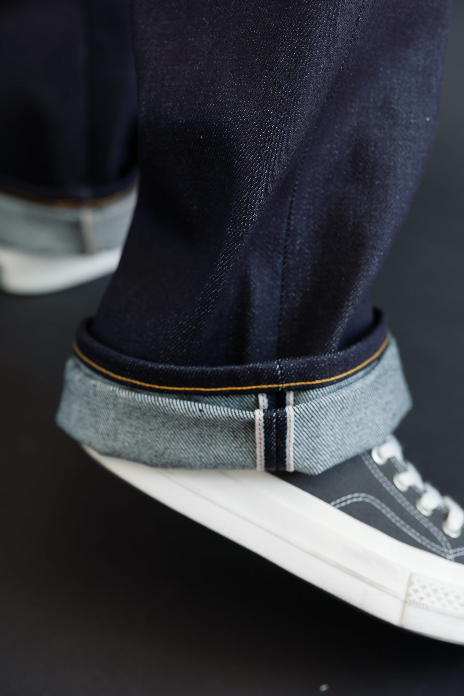 Close-up of the hem of dark indigo selvedge denim jeans worn by a male model, showing the light blue underside and orange stitching.