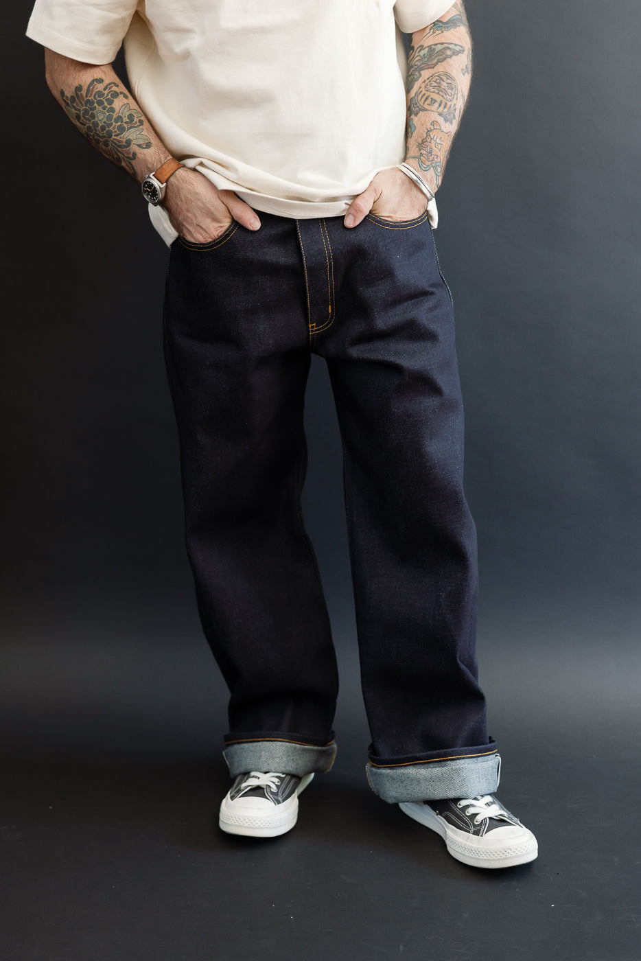 Full body front view of a male model with an athletic build wearing dark selvedge denim jeans and white sneakers.