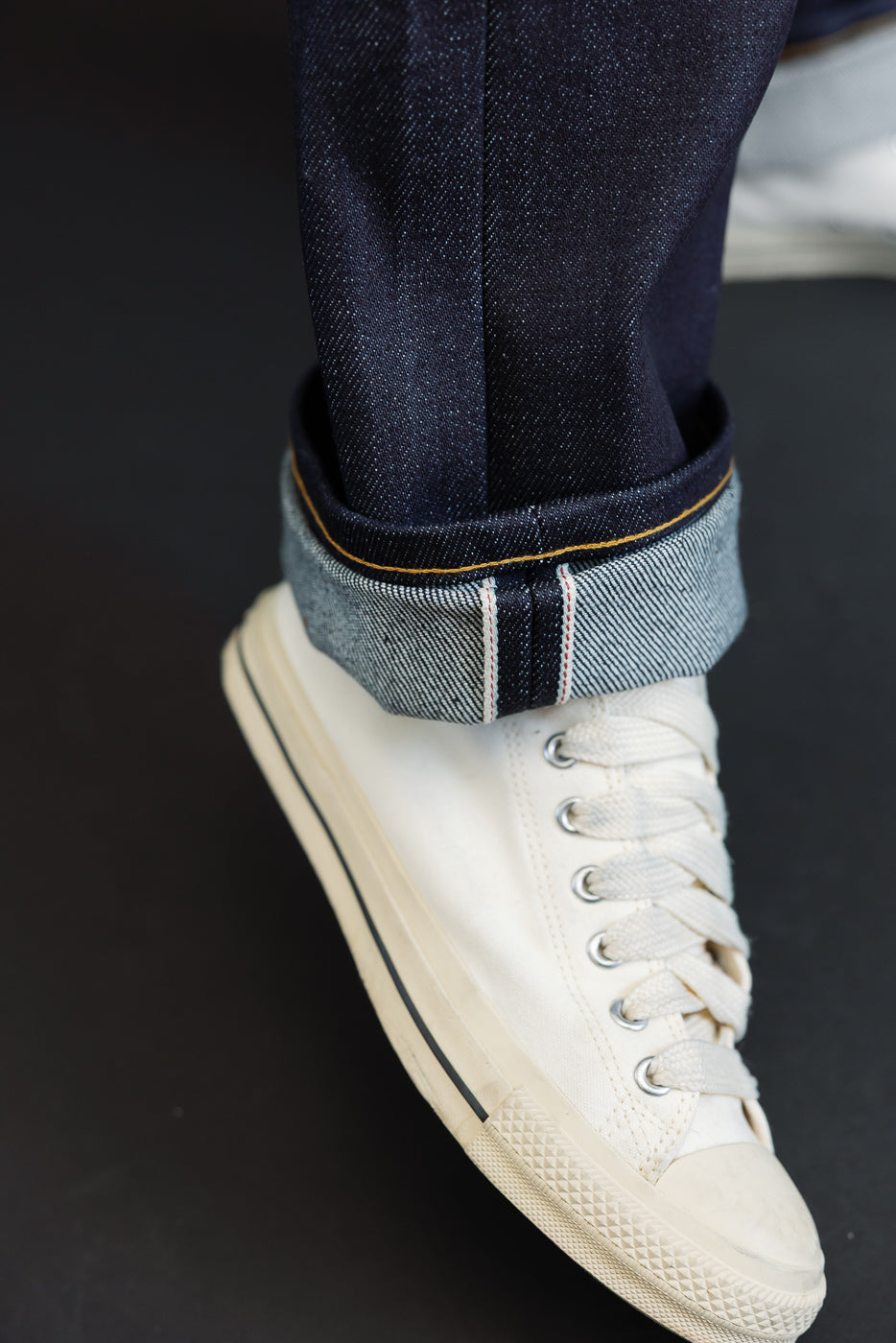Close-up of a male model's leg showing cuffed dark selvedge denim jeans and white canvas sneakers.
